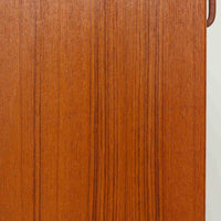 B.フリードホーゲンがデザインし、Bodafors社により製作された北欧ビンテージのディスプレイシェルフです。チーク材の美しい木目を活かした縦に伸びる端正なフレーム構成が印象的で、オープン棚と下部収納を組み合わせた実用性の高いデザインとなっています。上段は抜け感のある棚板構造で、書籍や器、オブジェを軽やかに飾ることができ、下段の引き戸収納は生活感の出やすい物をすっきりと収められます。脚付きによる軽快な佇まいと、経年変化によって深みを増した素材感が、空間に北欧ビンテージならではの落ち着いた存在感を与えます。