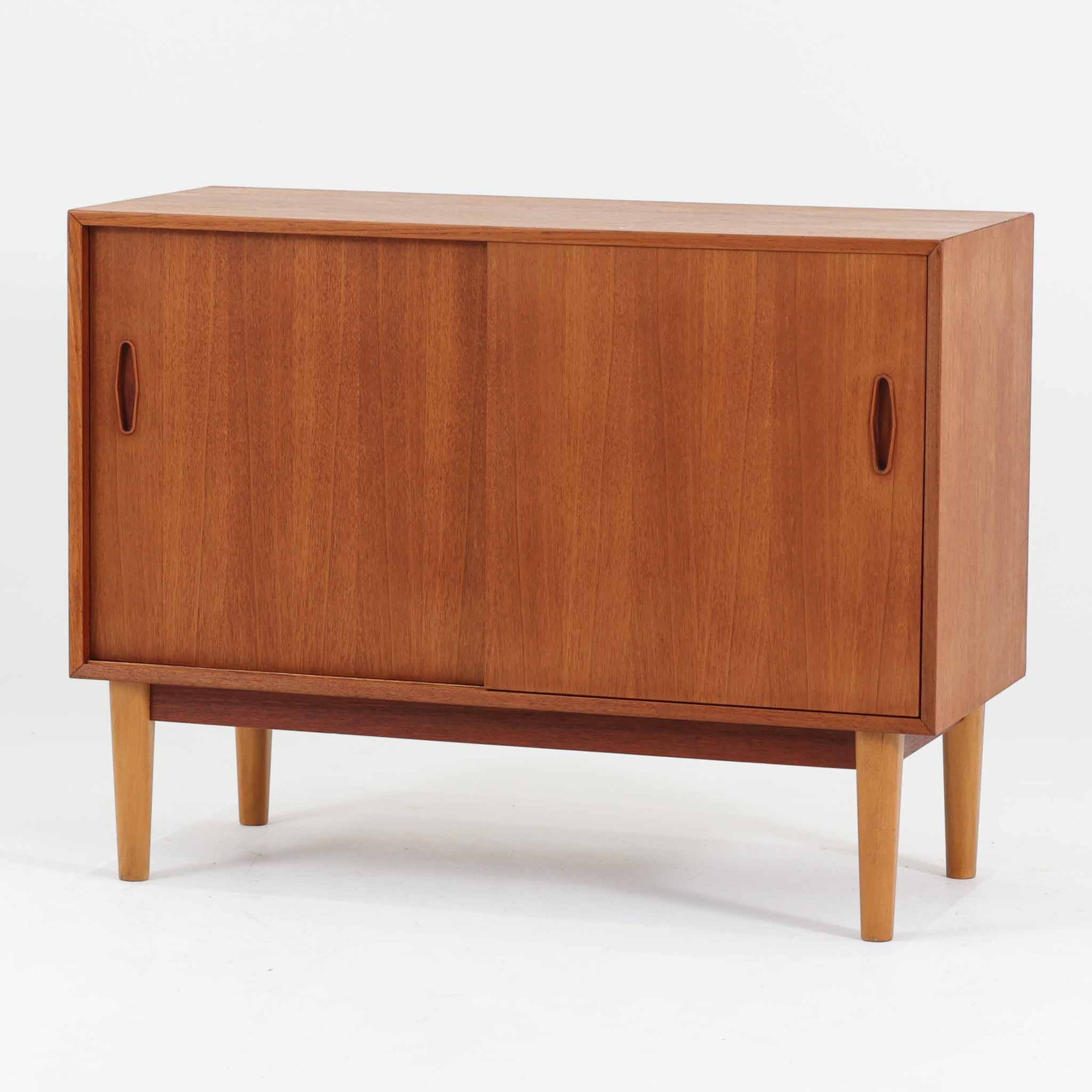 Nils Jonssonがデザインし、Hugo Troedsが製作した幅95cmの小ぶりなボードです。上質なチーク材を用いた端正な箱型フォルムに、楕円形の彫り込み取っ手がさりげないアクセントを添えています。引き戸仕様により限られた空間でも扱いやすく、内部には棚板を備え収納力も十分。すらりと伸びる脚部が軽やかな印象を与え、リビングや書斎、玄関先でも活躍します。経年により深みを増した木肌が北欧ビンテージならではの温もりと存在感を感じさせる一品です。