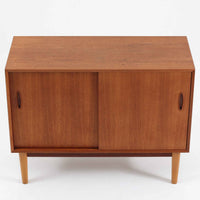 Nils Jonssonがデザインし、Hugo Troedsが製作した幅95cmの小ぶりなボードです。上質なチーク材を用いた端正な箱型フォルムに、楕円形の彫り込み取っ手がさりげないアクセントを添えています。引き戸仕様により限られた空間でも扱いやすく、内部には棚板を備え収納力も十分。すらりと伸びる脚部が軽やかな印象を与え、リビングや書斎、玄関先でも活躍します。経年により深みを増した木肌が北欧ビンテージならではの温もりと存在感を感じさせる一品です。