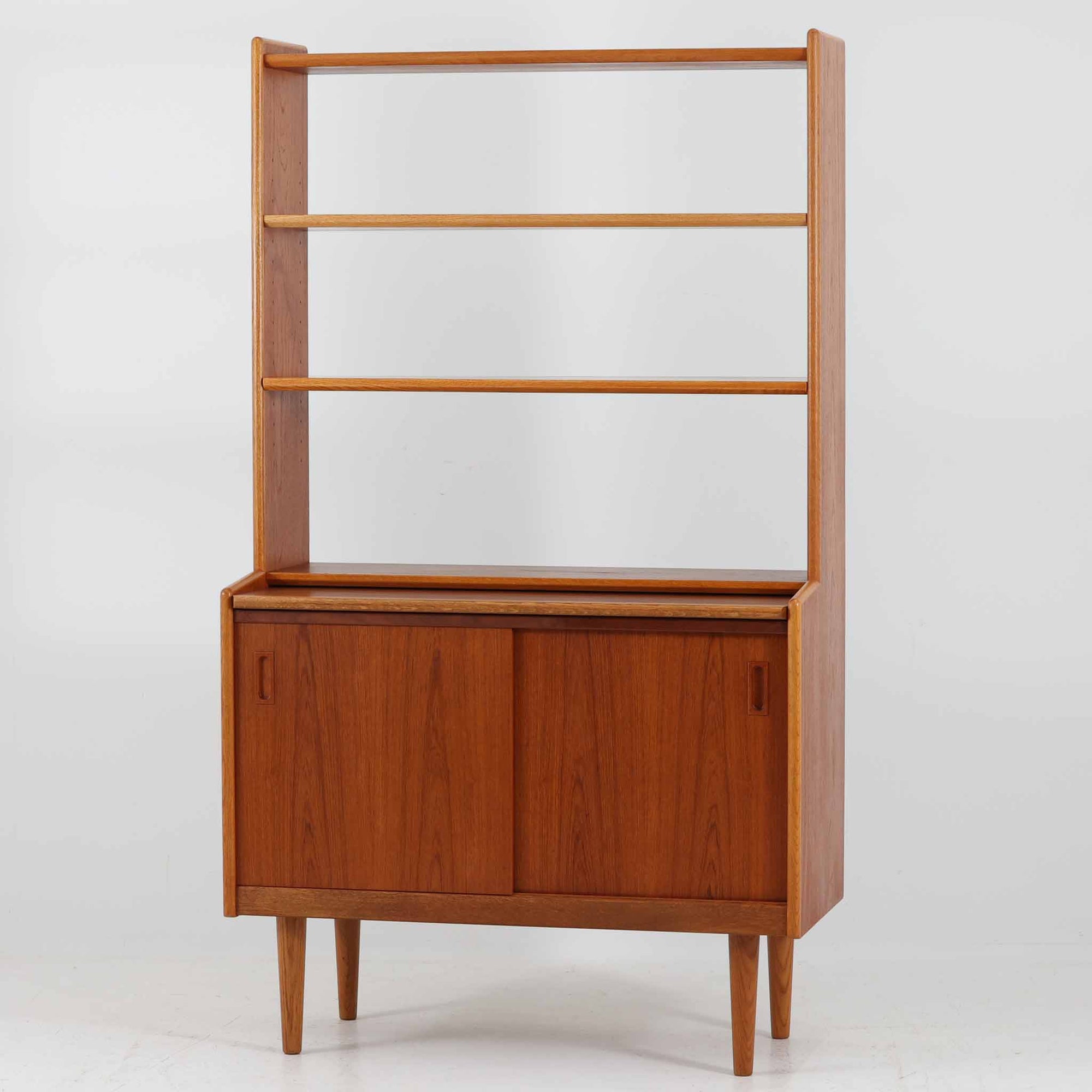 こちらのディスプレイシェルフは、チーク材とオーク材を組み合わせた柔らかなコントラストが魅力の北欧ビンテージ家具で、上部には奥行きの浅い棚板を2段備えた軽やかなディスプレイスペースに加えて天板部には引き棚が設けられており、雑貨や植物、本のレイアウトを楽しみながら作業スペースとしても活用でき、下部のスライド扉収納によって見せる収納と隠す収納を両立できるほか、細身の脚部が全体に浮遊感を与えて空間を圧迫しないバランスの良いデザインに仕上がっており、経年で深まった木肌の温もりも相まってリビングやダイニング、玄関周りにも取り入れやすい一台です。