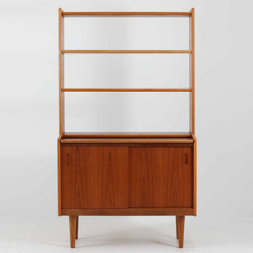 こちらのディスプレイシェルフは、チーク材とオーク材を組み合わせた柔らかなコントラストが魅力の北欧ビンテージ家具で、上部には奥行きの浅い棚板を2段備えた軽やかなディスプレイスペースに加えて天板部には引き棚が設けられており、雑貨や植物、本のレイアウトを楽しみながら作業スペースとしても活用でき、下部のスライド扉収納によって見せる収納と隠す収納を両立できるほか、細身の脚部が全体に浮遊感を与えて空間を圧迫しないバランスの良いデザインに仕上がっており、経年で深まった木肌の温もりも相まってリビングやダイニング、玄関周りにも取り入れやすい一台です。