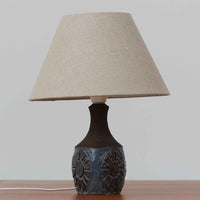 Chris HaslevがJetiのために手掛けたこちらのテーブルランプは、太陽を思わせるレリーフ模様をまとった陶器ベースと、素朴な土肌と釉薬の濃淡が織りなす豊かな表情が魅力で、コンパクトながら存在感を放つ北欧ビンテージ品です。上部のマットな質感が造形を引き締め、リネンシェード越しに広がるあたたかな灯りが空間をやさしく包み込み、リビングやベッドサイドのアクセントとして取り入れやすい一台に仕上がっています。