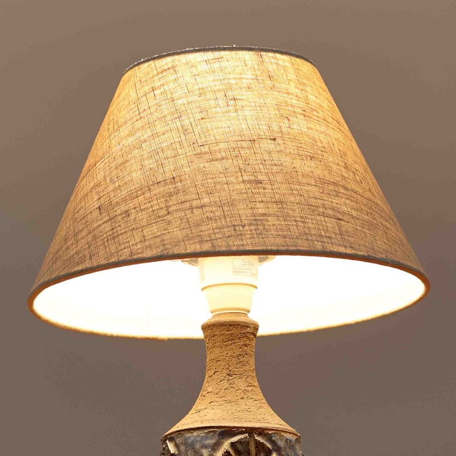 Chris HaslevがJetiのために手掛けたこちらのテーブルランプは、太陽を思わせるレリーフ模様をまとった陶器ベースと、素朴な土肌と釉薬の濃淡が織りなす豊かな表情が魅力で、コンパクトながら存在感を放つ北欧ビンテージ品です。上部のマットな質感が造形を引き締め、リネンシェード越しに広がるあたたかな灯りが空間をやさしく包み込み、リビングやベッドサイドのアクセントとして取り入れやすい一台に仕上がっています。