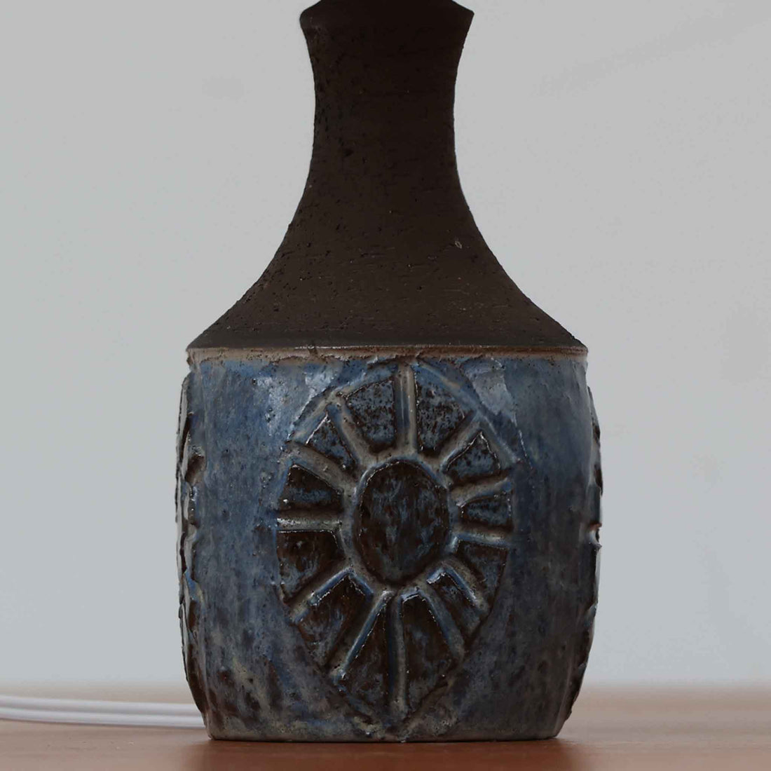 Chris HaslevがJetiのために手掛けたこちらのテーブルランプは、太陽を思わせるレリーフ模様をまとった陶器ベースと、素朴な土肌と釉薬の濃淡が織りなす豊かな表情が魅力で、コンパクトながら存在感を放つ北欧ビンテージ品です。上部のマットな質感が造形を引き締め、リネンシェード越しに広がるあたたかな灯りが空間をやさしく包み込み、リビングやベッドサイドのアクセントとして取り入れやすい一台に仕上がっています。