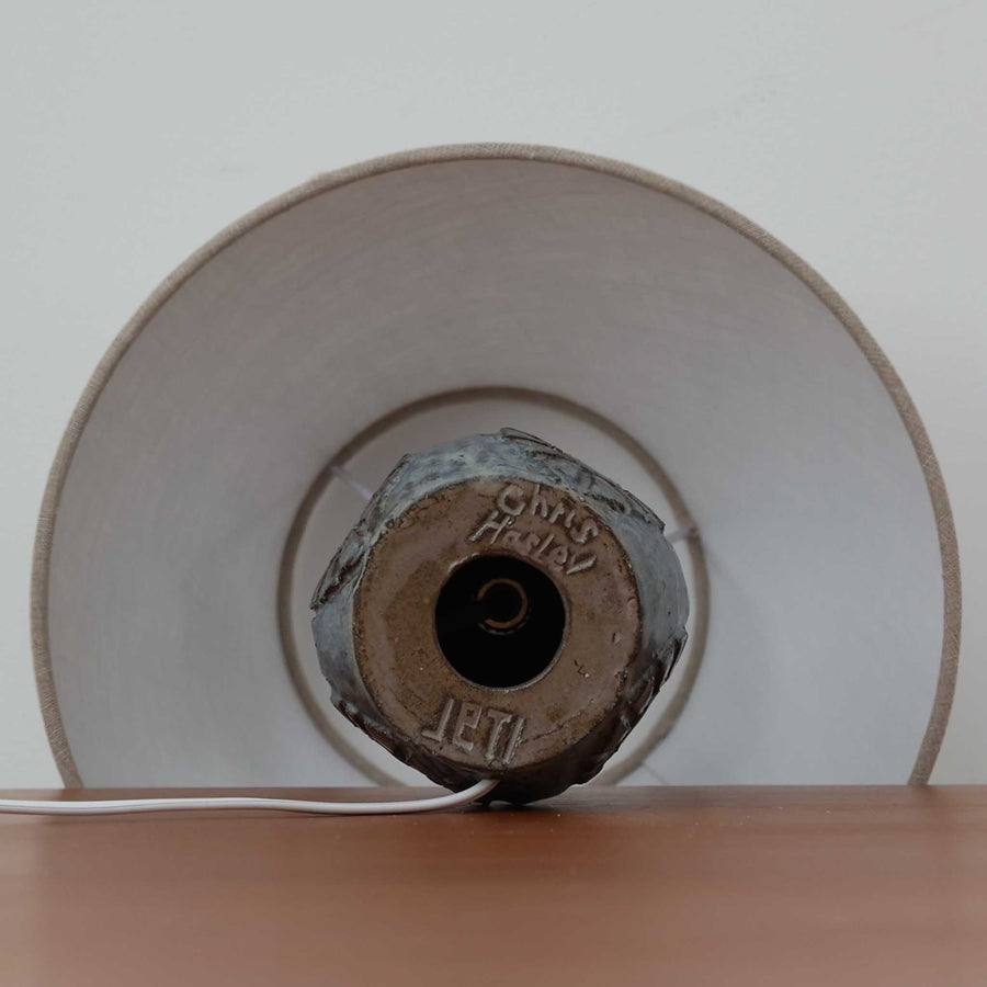 Chris HaslevがJetiのために手掛けたこちらのテーブルランプは、太陽を思わせるレリーフ模様をまとった陶器ベースと、素朴な土肌と釉薬の濃淡が織りなす豊かな表情が魅力で、コンパクトながら存在感を放つ北欧ビンテージ品です。上部のマットな質感が造形を引き締め、リネンシェード越しに広がるあたたかな灯りが空間をやさしく包み込み、リビングやベッドサイドのアクセントとして取り入れやすい一台に仕上がっています。