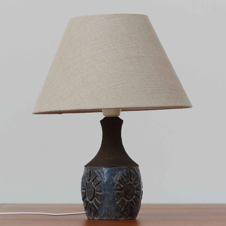 Chris HaslevがJetiのために手掛けたこちらのテーブルランプは、太陽を思わせるレリーフ模様をまとった陶器ベースと、素朴な土肌と釉薬の濃淡が織りなす豊かな表情が魅力で、コンパクトながら存在感を放つ北欧ビンテージ品です。上部のマットな質感が造形を引き締め、リネンシェード越しに広がるあたたかな灯りが空間をやさしく包み込み、リビングやベッドサイドのアクセントとして取り入れやすい一台に仕上がっています。