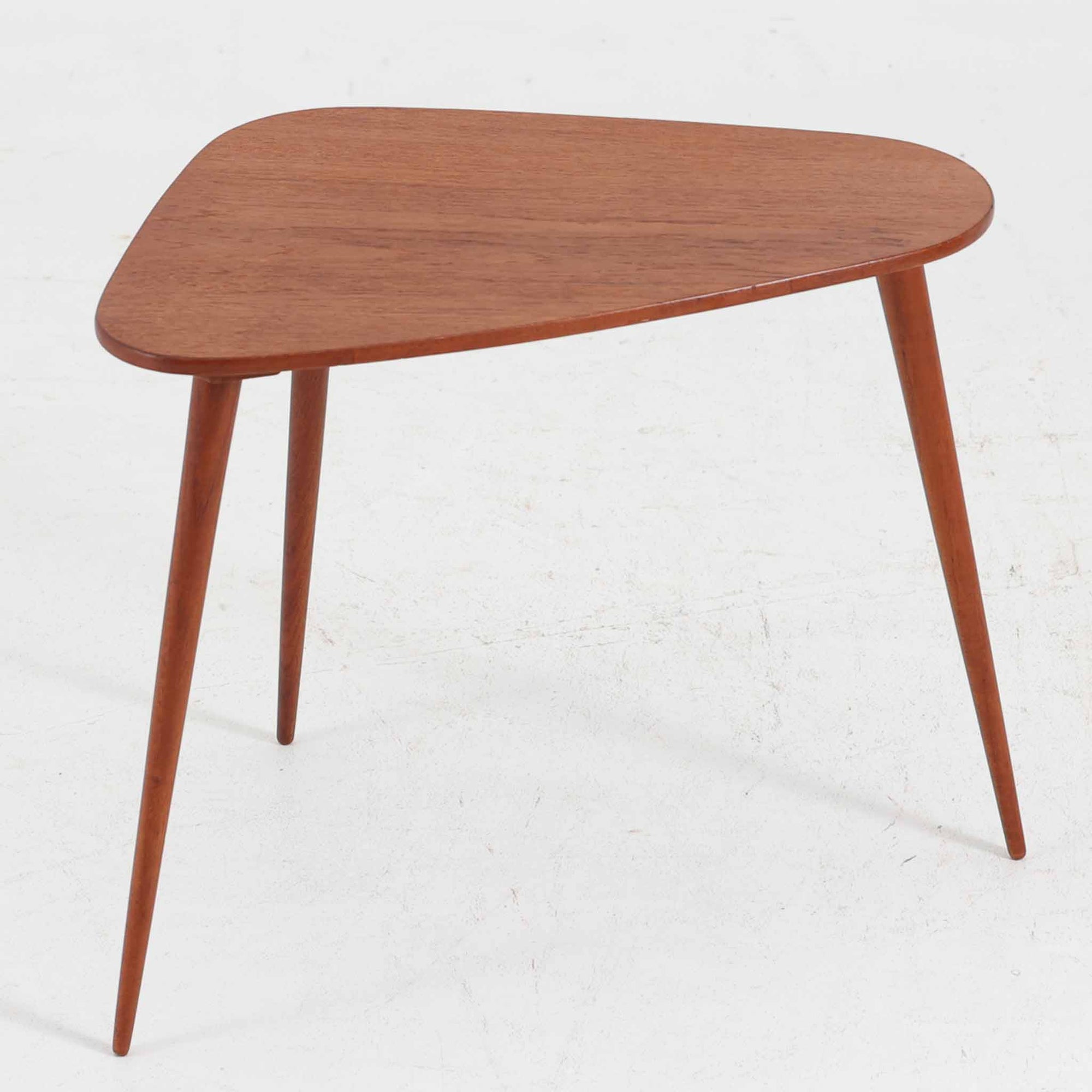 デンマーク製のチーク材サイドテーブルです。三角形に近い柔らかな曲線を描く天板と、すらりと伸びた3本脚が印象的なデザインで、ミッドセンチュリーらしい軽快な佇まいが魅力です。職人による丁寧な削り出しが際立ち、チーク材特有の深みある色合いと滑らかな手触りが楽しめます。コンパクトながら存在感があり、ソファ横やベッドサイドの小物置きとしても最適な一台です。