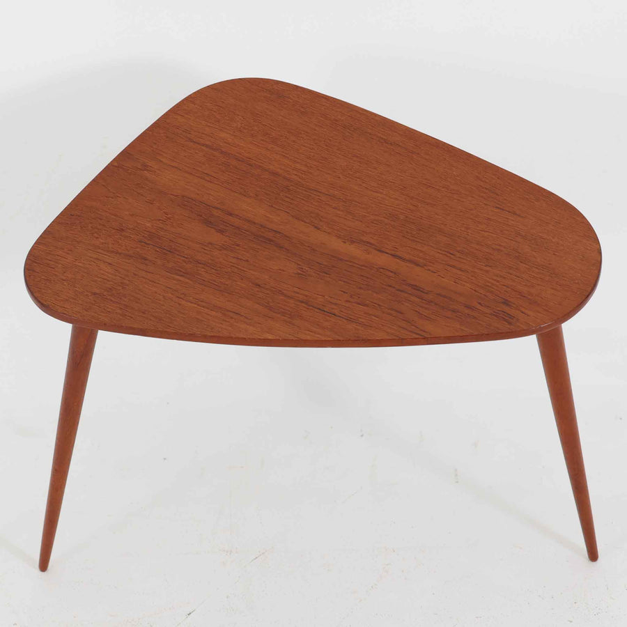 デンマーク製のチーク材サイドテーブルです。三角形に近い柔らかな曲線を描く天板と、すらりと伸びた3本脚が印象的なデザインで、ミッドセンチュリーらしい軽快な佇まいが魅力です。職人による丁寧な削り出しが際立ち、チーク材特有の深みある色合いと滑らかな手触りが楽しめます。コンパクトながら存在感があり、ソファ横やベッドサイドの小物置きとしても最適な一台です。