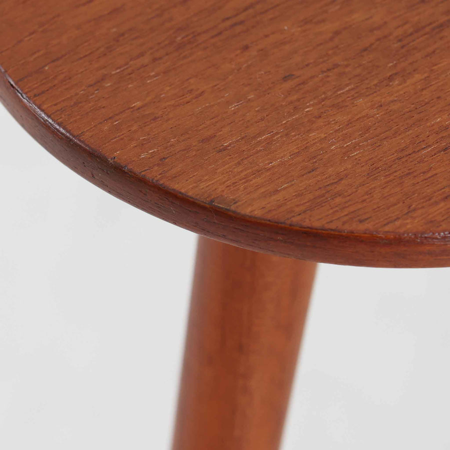 デンマーク製のチーク材サイドテーブルです。三角形に近い柔らかな曲線を描く天板と、すらりと伸びた3本脚が印象的なデザインで、ミッドセンチュリーらしい軽快な佇まいが魅力です。職人による丁寧な削り出しが際立ち、チーク材特有の深みある色合いと滑らかな手触りが楽しめます。コンパクトながら存在感があり、ソファ横やベッドサイドの小物置きとしても最適な一台です。