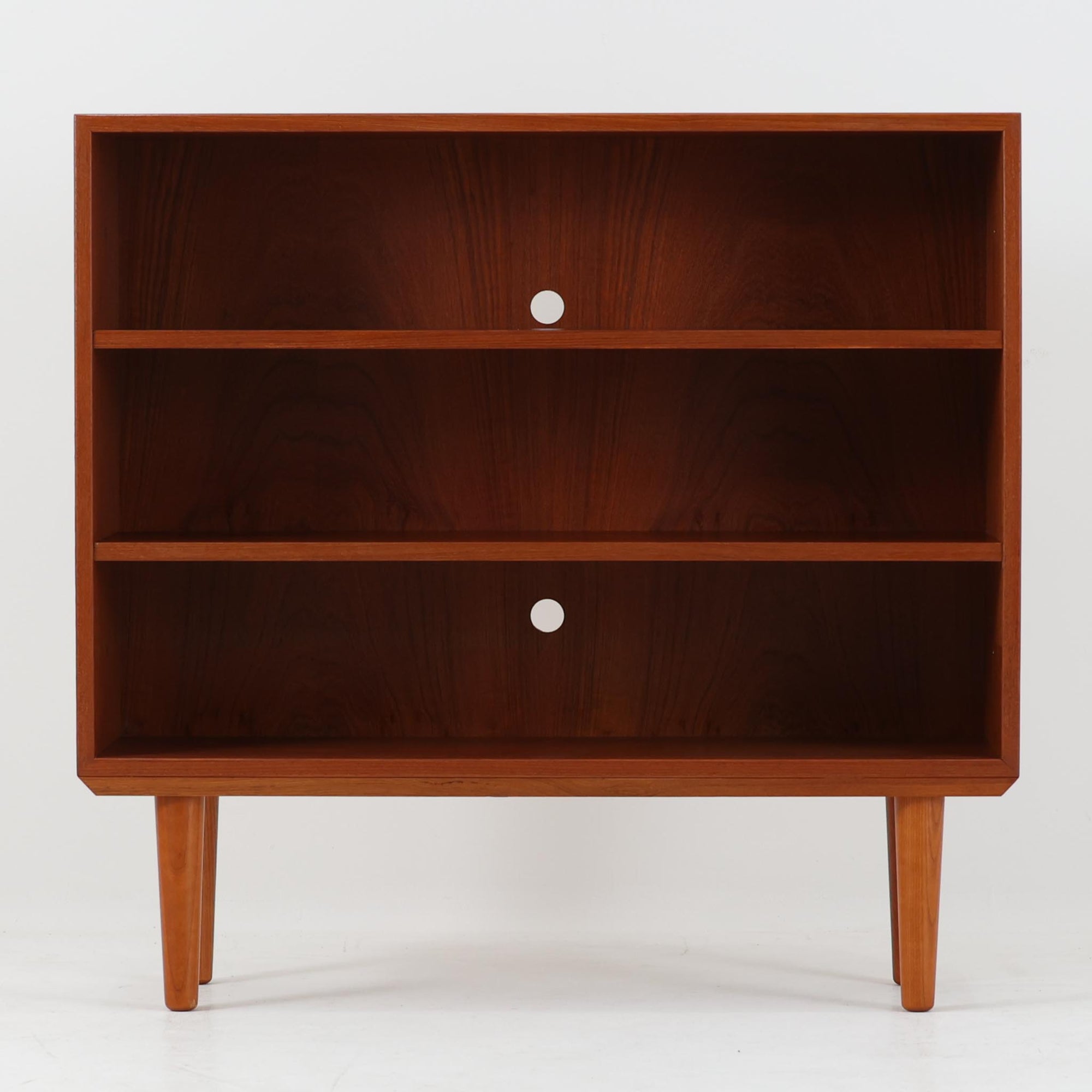 Hjornebo Mobelfabrik（ヒョルネボ・モーベルファブリック）によるデンマーク製のチーク材ブックシェルフです。幅86cmの使いやすいサイズに、すっきりとしたオープン構造と丸みを帯びた脚部を組み合わせた、軽やかな北欧デザインが魅力の一台。内部は2枚の棚板で実用的に仕切られ、書籍や雑誌の収納はもちろん、オブジェや器、オーディオ機器を飾るディスプレイシェルフとしても美しく映えます。背板には配線穴が設けられており、AVボードやサイドボード感覚でも取り入れやすい設計。経年により深みを増したチークの美しい木目と丁寧な造りが、ビンテージならではの上質な存在感と希少性を空間にもたらします。