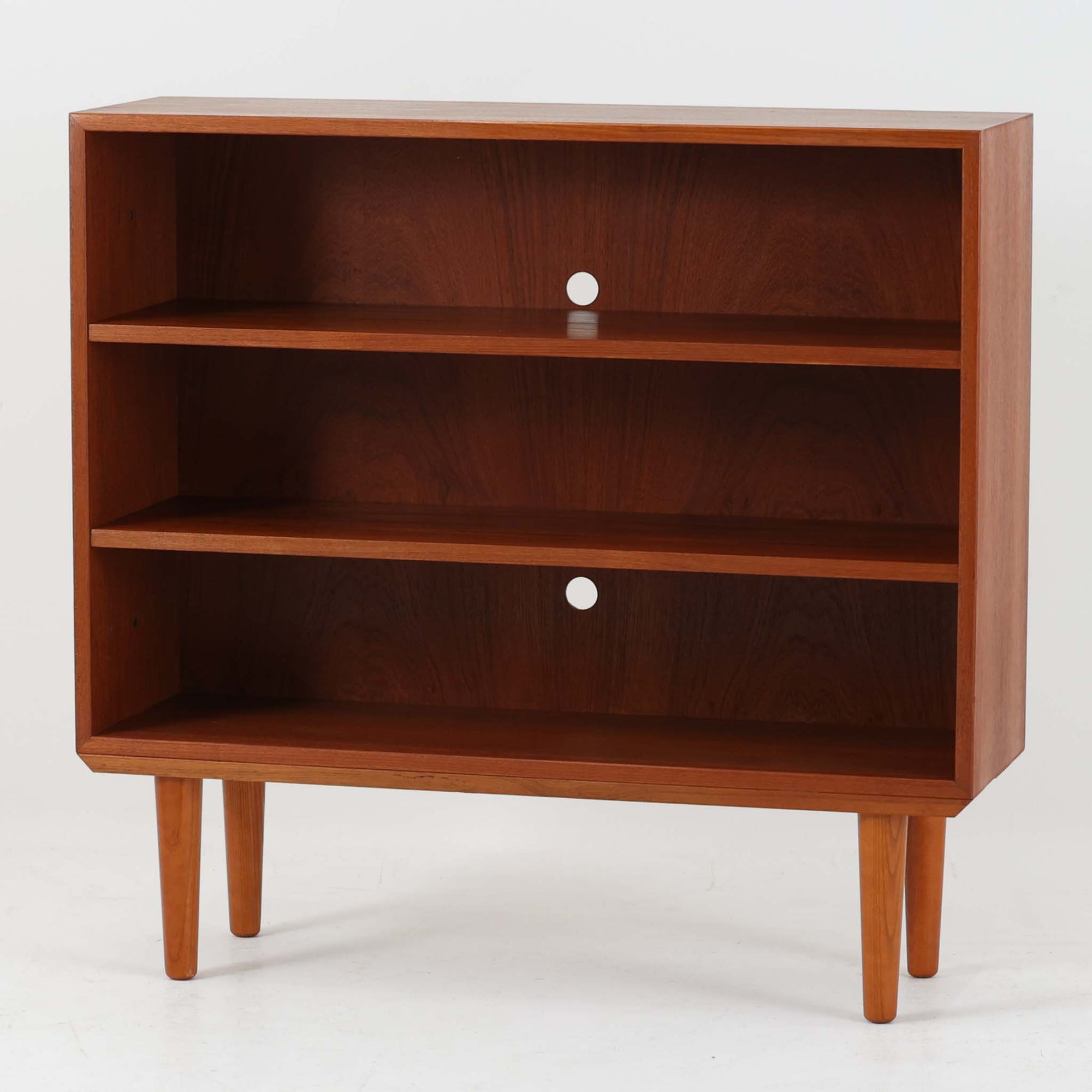 Hjornebo Mobelfabrik（ヒョルネボ・モーベルファブリック）によるデンマーク製のチーク材ブックシェルフです。幅86cmの使いやすいサイズに、すっきりとしたオープン構造と丸みを帯びた脚部を組み合わせた、軽やかな北欧デザインが魅力の一台。内部は2枚の棚板で実用的に仕切られ、書籍や雑誌の収納はもちろん、オブジェや器、オーディオ機器を飾るディスプレイシェルフとしても美しく映えます。背板には配線穴が設けられており、AVボードやサイドボード感覚でも取り入れやすい設計。経年により深みを増したチークの美しい木目と丁寧な造りが、ビンテージならではの上質な存在感と希少性を空間にもたらします。