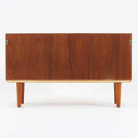 デンマークで製作されたチーク材のローボードです。幅100cmの小ぶりなサイズながら、温かみのある木目とシンプルな直線的フォルムが北欧らしい落ち着いた佇まいを演出します。前面には手掛け付きのスライド扉が備えられ、内部収納をすっきりと隠しながら使いやすさも確保。細身の脚部が軽やかな印象を与え、リビングや玄関、ベッドサイドなど様々な空間で活躍します。背板には壁掛け用の金具が取り付けられており、収納内部にはDIYによる木材の追加や小さな穴が見られますが、ビンテージ品ならではの味わいとして楽しめる一台です。