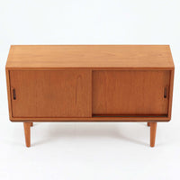 デンマークで製作されたチーク材のローボードです。幅100cmの小ぶりなサイズながら、温かみのある木目とシンプルな直線的フォルムが北欧らしい落ち着いた佇まいを演出します。前面には手掛け付きのスライド扉が備えられ、内部収納をすっきりと隠しながら使いやすさも確保。細身の脚部が軽やかな印象を与え、リビングや玄関、ベッドサイドなど様々な空間で活躍します。背板には壁掛け用の金具が取り付けられており、収納内部にはDIYによる木材の追加や小さな穴が見られますが、ビンテージ品ならではの味わいとして楽しめる一台です。