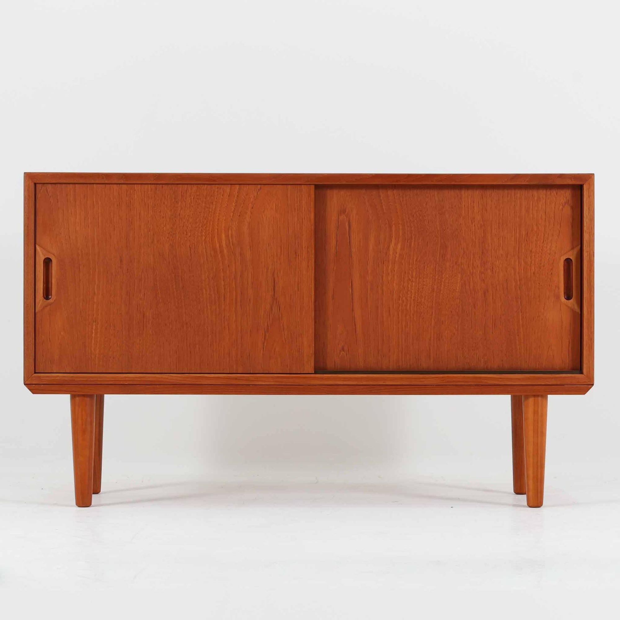 デンマークで製作されたチーク材のローボードです。幅100cmの小ぶりなサイズながら、温かみのある木目とシンプルな直線的フォルムが北欧らしい落ち着いた佇まいを演出します。前面には手掛け付きのスライド扉が備えられ、内部収納をすっきりと隠しながら使いやすさも確保。細身の脚部が軽やかな印象を与え、リビングや玄関、ベッドサイドなど様々な空間で活躍します。背板には壁掛け用の金具が取り付けられており、収納内部にはDIYによる木材の追加や小さな穴が見られますが、ビンテージ品ならではの味わいとして楽しめる一台です。