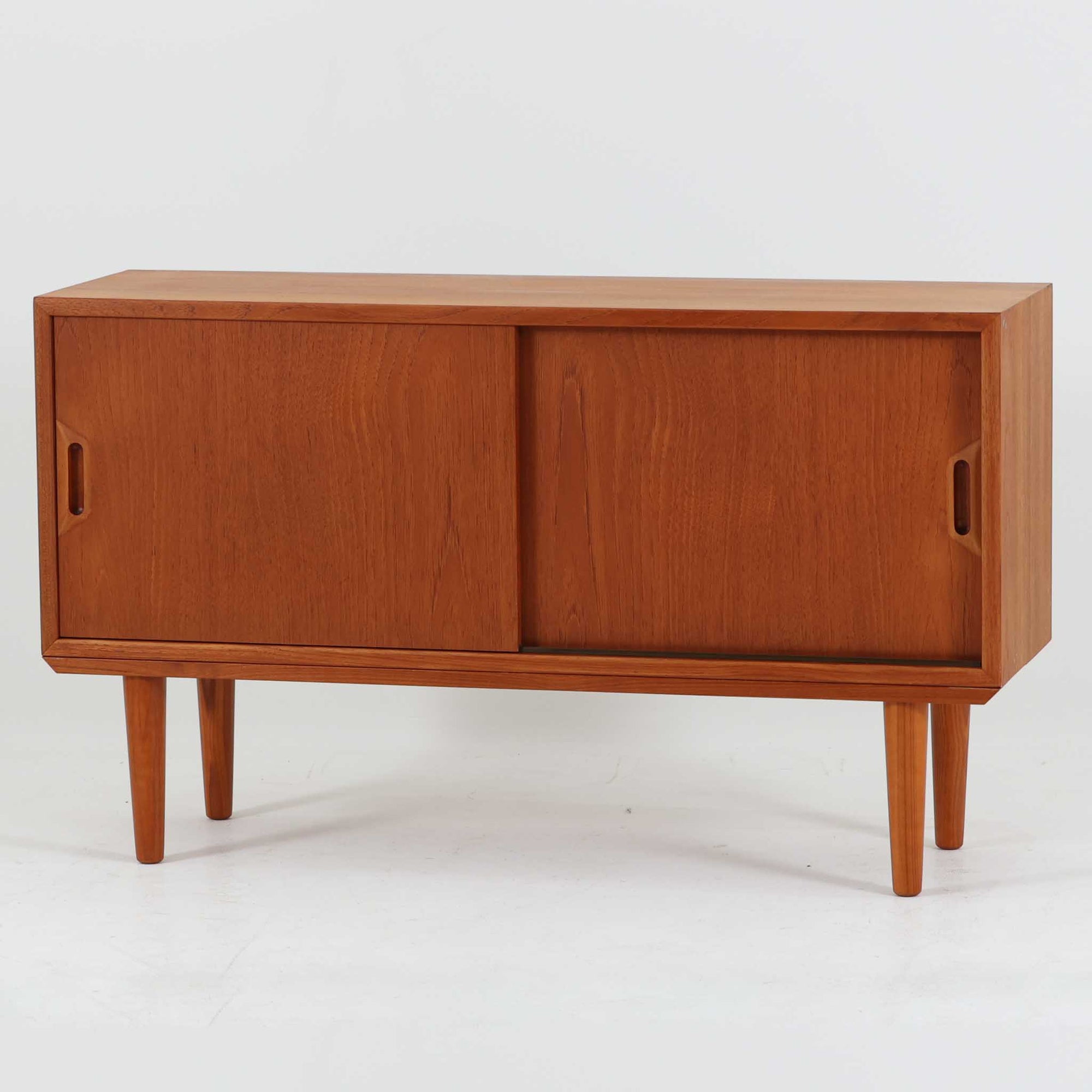 デンマークで製作されたチーク材のローボードです。幅100cmの小ぶりなサイズながら、温かみのある木目とシンプルな直線的フォルムが北欧らしい落ち着いた佇まいを演出します。前面には手掛け付きのスライド扉が備えられ、内部収納をすっきりと隠しながら使いやすさも確保。細身の脚部が軽やかな印象を与え、リビングや玄関、ベッドサイドなど様々な空間で活躍します。背板には壁掛け用の金具が取り付けられており、収納内部にはDIYによる木材の追加や小さな穴が見られますが、ビンテージ品ならではの味わいとして楽しめる一台です。