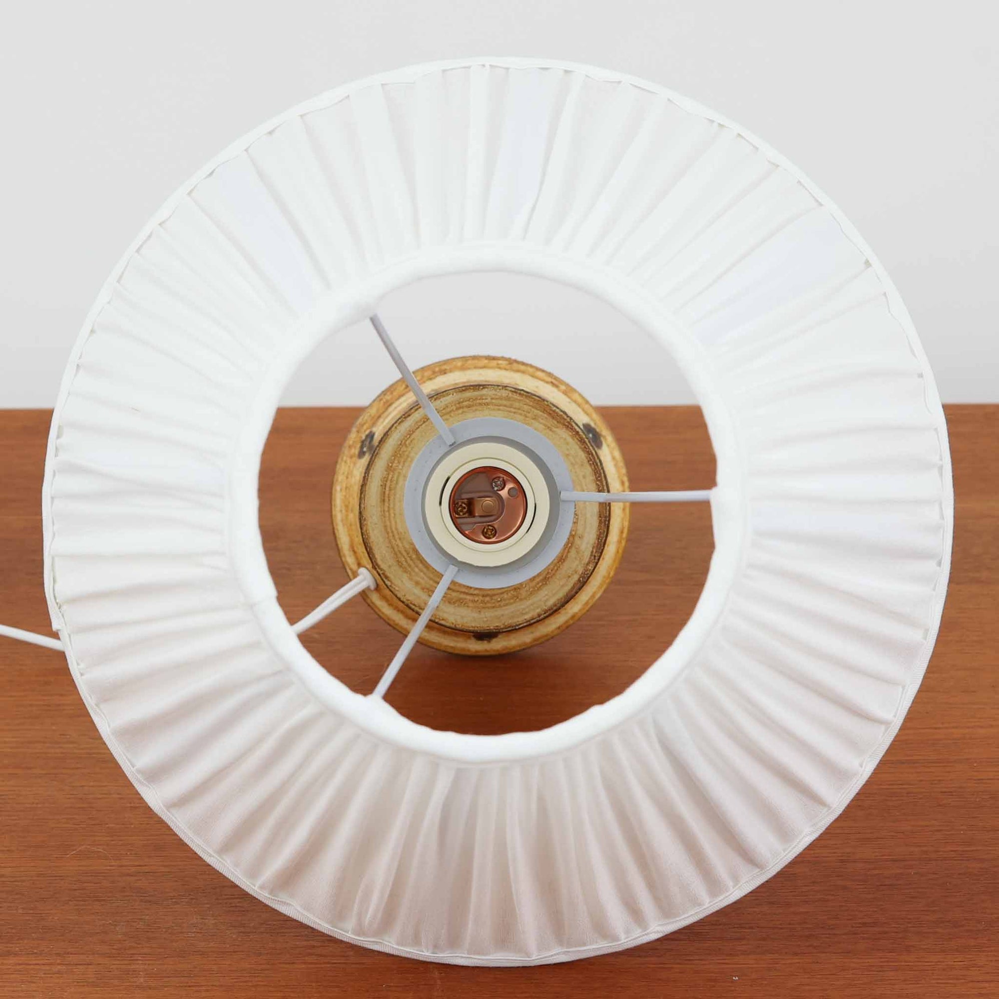 Bartholdy Keramikが手掛けたデンマーク製テーブルランプです。高さ40cmの程よいサイズ感に、陶磁器の素朴な土味とプリーツ状のリネンシェードが美しく調和した北欧ビンテージらしい一台。ベースには花火や草花を思わせる放射状の絵付けが施され、手仕事ならではの柔らかな揺らぎと釉薬の景色が豊かな表情を生み出しています。ややくびれを持たせたシルエットが軽やかな印象を与え、点灯時にはシェード越しの温かな光が絵柄をやさしく引き立て、空間に穏やかなアクセントを添えます。リビングやベッドサイド、チェスト上で北欧陶器ならではの温もりと希少性を楽しめる、存在感あるビンテージ照明です。