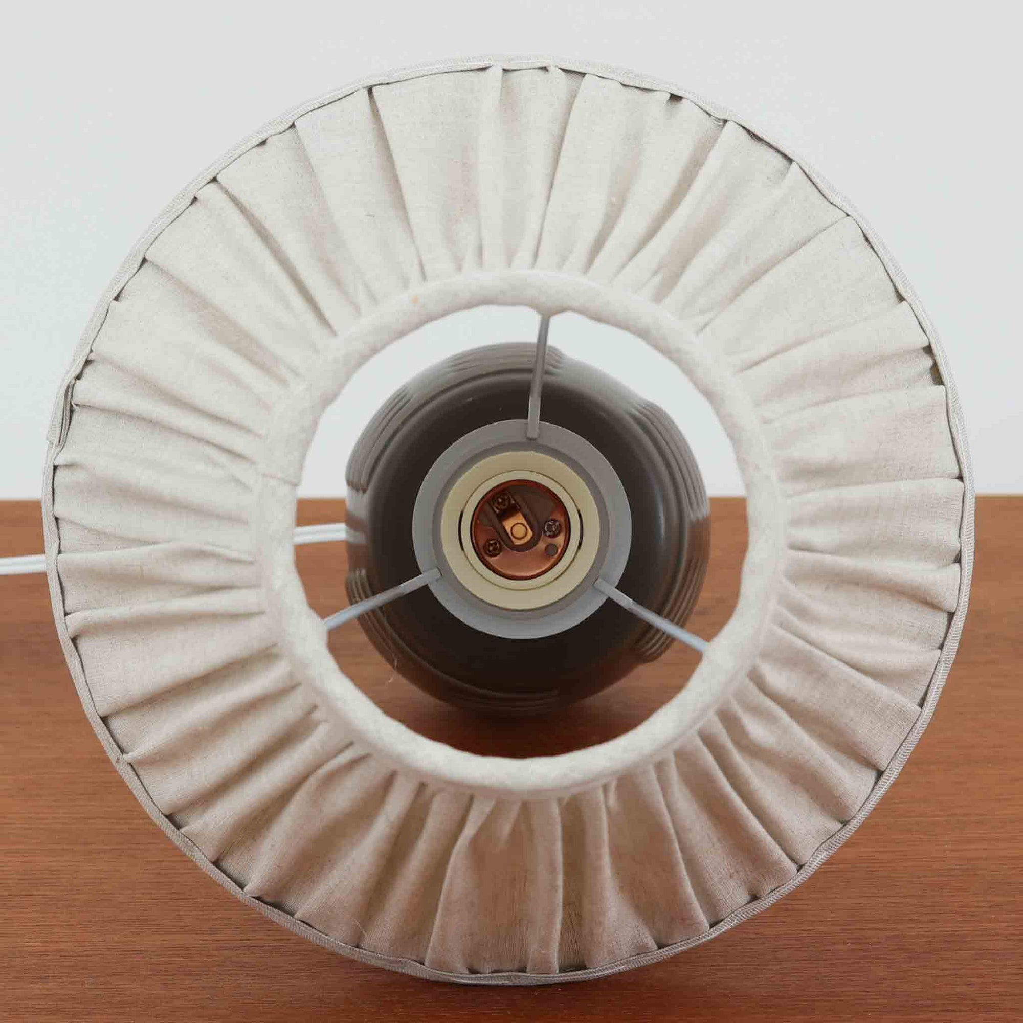 Rigmor Nielsenがデザインし、Soholm Keramikが手掛けたテーブルランプです。高さ37cmの取り入れやすいサイズに、艶やかな陶磁器ベースとナチュラルなリネンシェードを組み合わせた、落ち着きある北欧ビンテージらしい一台。ベースには規則的な立体リブ装飾が施され、陰影豊かな表情と彫刻的な存在感を生み出しています。点灯時にはシェード越しの柔らかな光が釉薬の奥行きや貫入の表情を引き立て、チェストやサイドテーブル、ベッドサイドに穏やかな雰囲気を演出。Rigmor Nielsenらしい造形美とSoholmの高い陶芸技術が感じられる、希少性の高いビンテージ照明です。