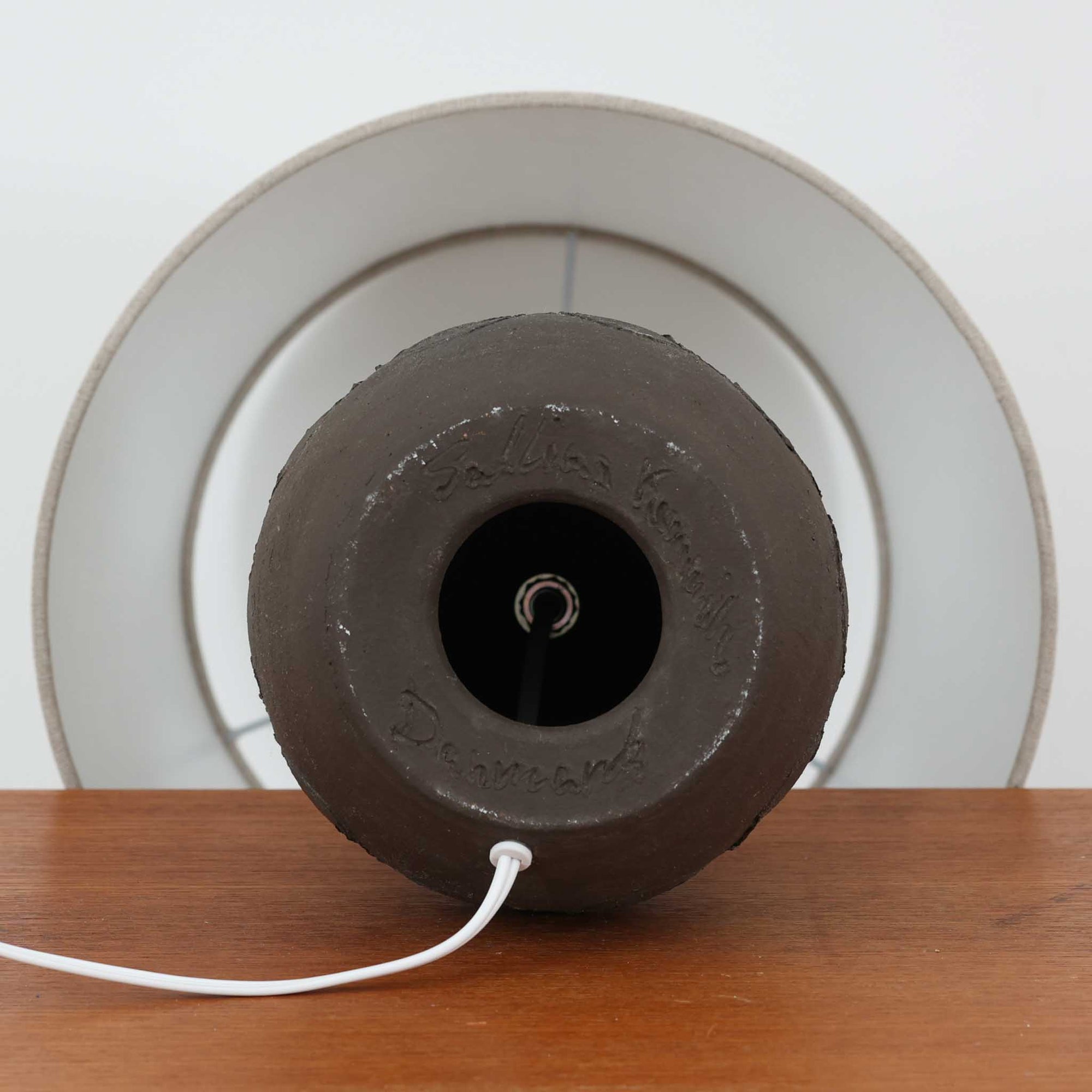 Salling Keramik製のデンマーク製テーブルランプです。陶磁器ベースは、落ち着いたダークブラウンのマットな質感と、上部にわずかな艶を残した焼き締めのような表情が魅力で、丸みのある壺型フォルムにくびれたネックを組み合わせたバランスの良い造形が印象的です。胴部には連続する円環モチーフのレリーフ装飾が巡らされ、素朴さの中に北欧らしいモダンなリズムを添えています。リネンシェードを通した柔らかな灯りが土味豊かな陶肌をやさしく引き立て、サイドテーブルやチェスト上、ベッドサイドの間接照明として使いやすいサイズ感。経年により深みを増した陶器の風合いと職人の手仕事を感じる表面の揺らぎが、ビンテージならではの温もりと希少性を高めています。
