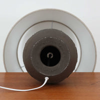 Salling Keramik製のデンマーク製テーブルランプです。陶磁器ベースは、落ち着いたダークブラウンのマットな質感と、上部にわずかな艶を残した焼き締めのような表情が魅力で、丸みのある壺型フォルムにくびれたネックを組み合わせたバランスの良い造形が印象的です。胴部には連続する円環モチーフのレリーフ装飾が巡らされ、素朴さの中に北欧らしいモダンなリズムを添えています。リネンシェードを通した柔らかな灯りが土味豊かな陶肌をやさしく引き立て、サイドテーブルやチェスト上、ベッドサイドの間接照明として使いやすいサイズ感。経年により深みを増した陶器の風合いと職人の手仕事を感じる表面の揺らぎが、ビンテージならではの温もりと希少性を高めています。