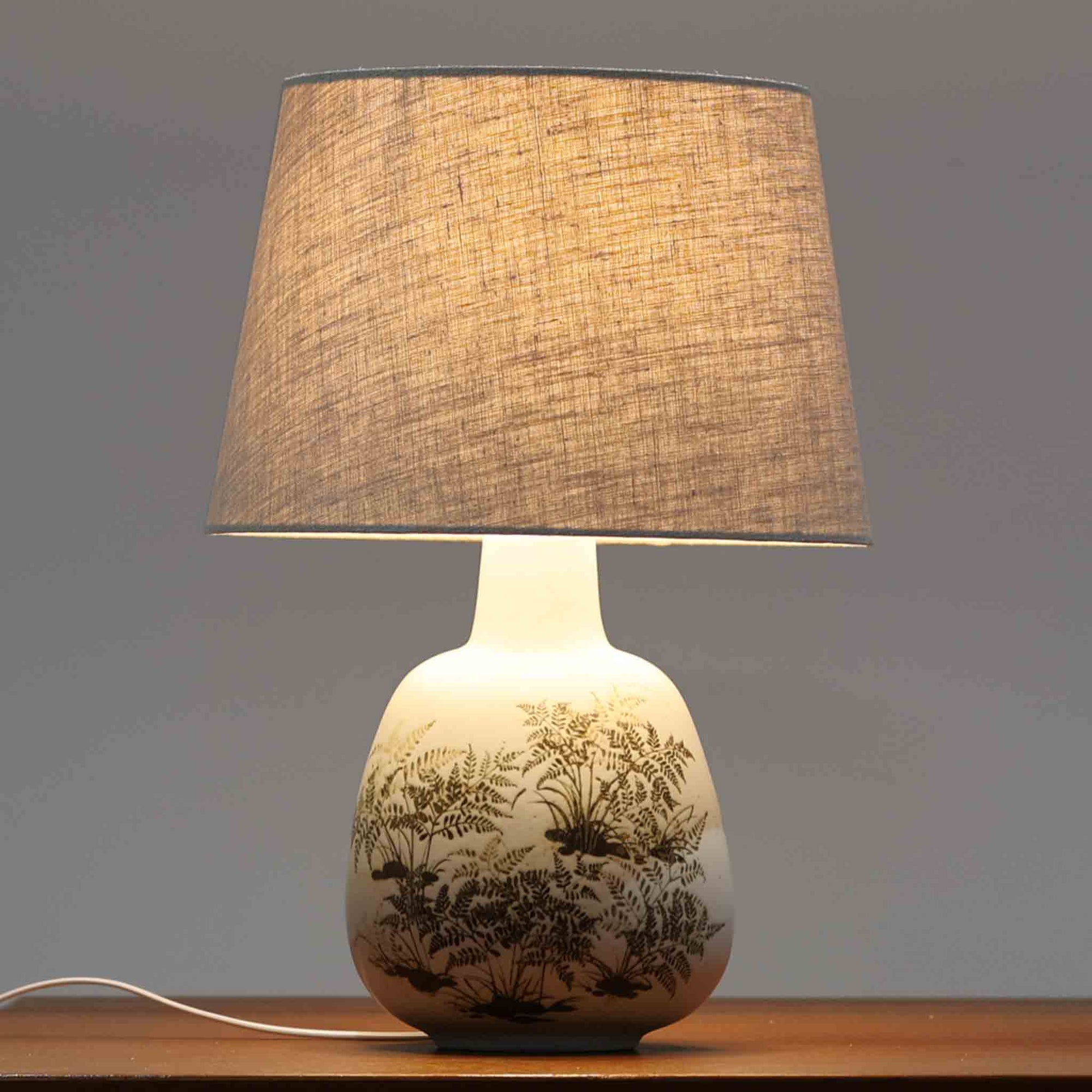 Royal CopenhagenのDIANAシリーズ、Nils Thorssonデザインによるデンマーク製ビンテージテーブルランプです。丸みを帯びた陶磁器ベースに繊細なシダ植物のモチーフが丁寧に描かれ、白磁の柔らかな余白とブラウンの絵付けが上品で静かな存在感を生み出します。ニルス・トーソンらしい詩情ある図案と、ロイヤルコペンハーゲンならではの高い絵付け技術が際立つ希少なモデル。リネンシェード越しのやわらかな灯りが絵柄を美しく浮かび上がらせ、ベッドサイドやチェスト、リビングのアクセント照明として空間に落ち着いた北欧らしさを添えてくれます。