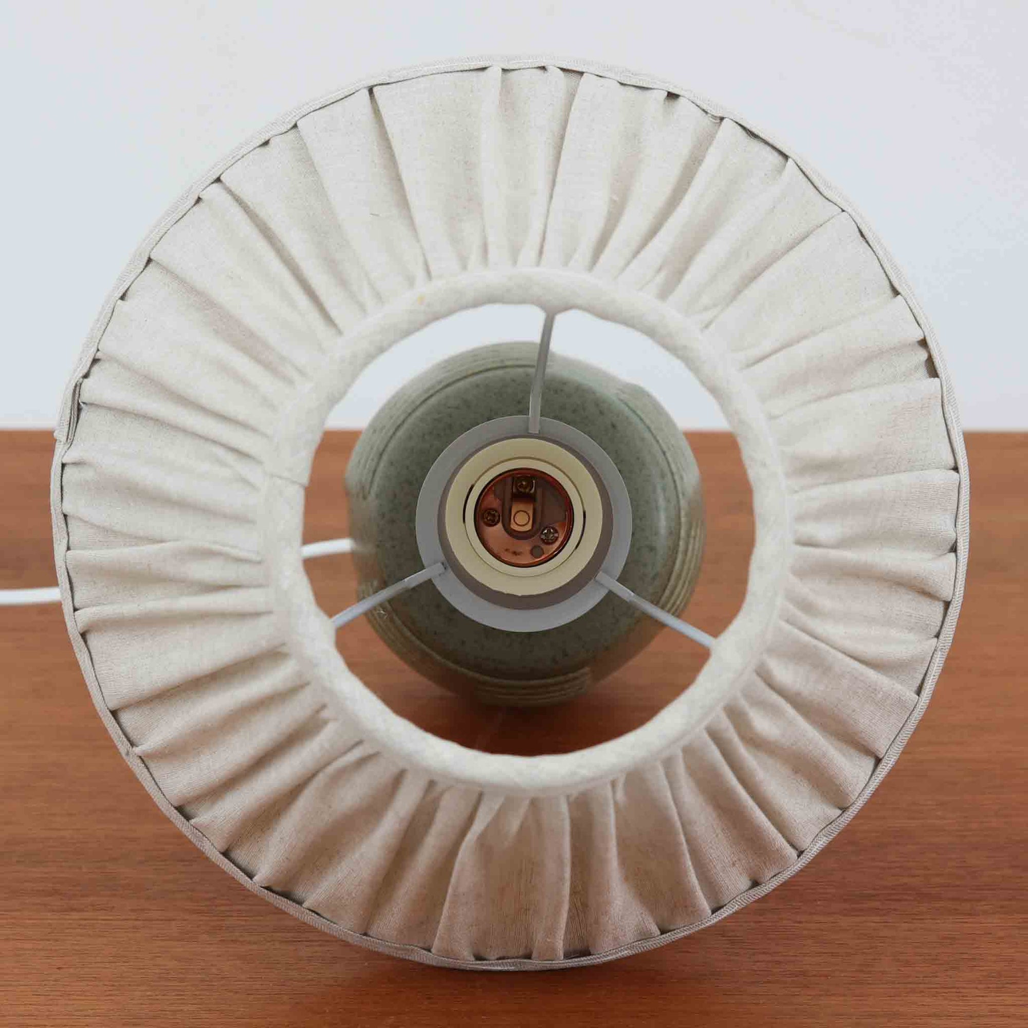 Einar Johansenがデザインし、Soholm Keramikが手掛けたテーブルランプです。高さ37cmの取り入れやすいサイズに、艶やかな陶磁器ベースとナチュラルなリネンシェードを組み合わせた、落ち着きある北欧ビンテージらしい一台。ベースには規則的な立体リブ装飾が施され、陰影豊かな表情と彫刻的な存在感を生み出しています。点灯時にはシェード越しの柔らかな光が釉薬の奥行きや貫入の表情を引き立て、チェストやサイドテーブル、ベッドサイドに穏やかな雰囲気を演出。Einar Johansenらしい造形美とSoholmの高い陶芸技術が感じられる、希少性の高いビンテージ照明です。