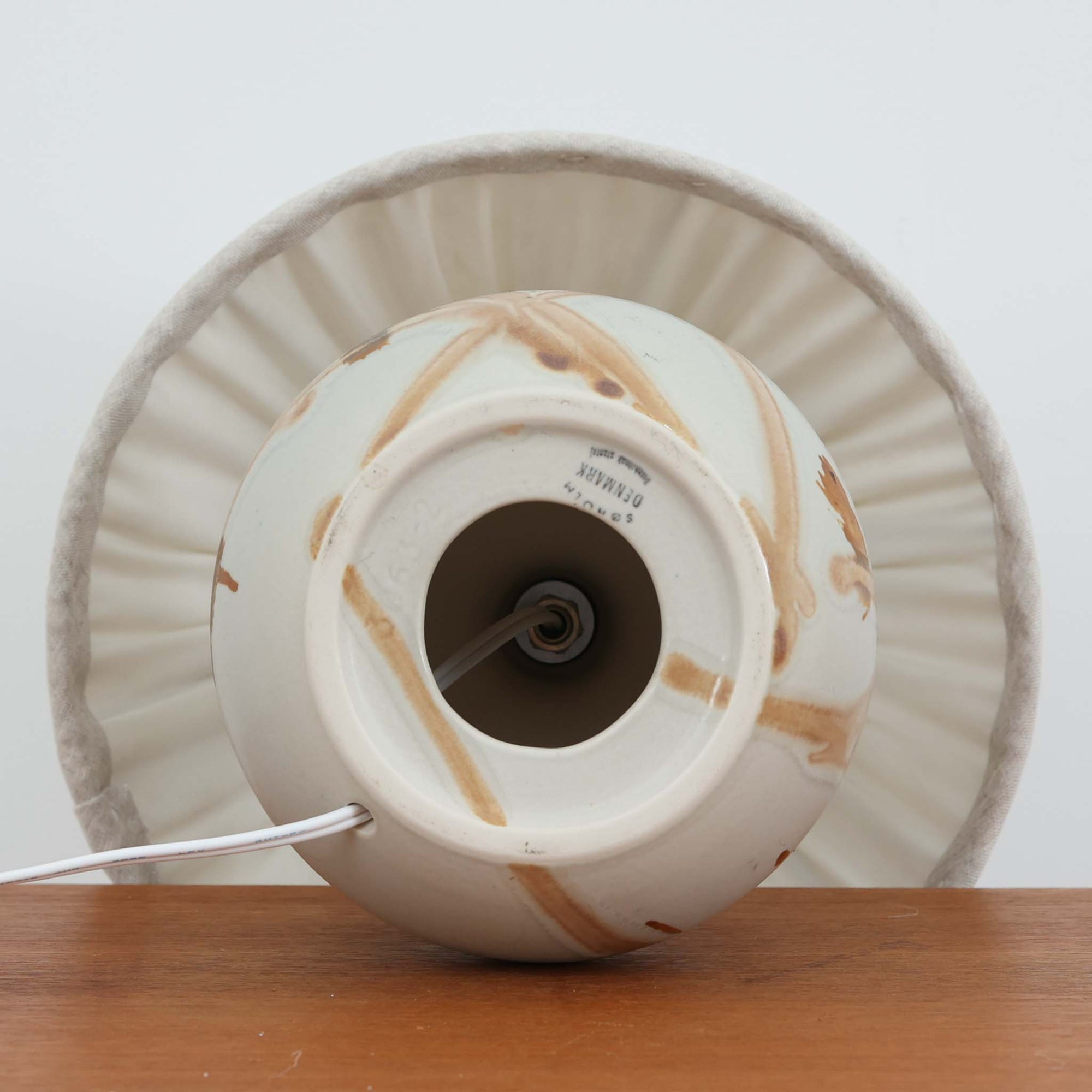 Soholm Keramikのモデル3063-2、Noomi Backhausenデザインによるデンマーク製ビンテージテーブルランプです。丸みを帯びた愛らしい陶磁器ベースに、ノーミ・バックハウゼンらしい抽象的で有機的なブラウンの流線模様が描かれ、柔らかなアイボリー地とのコントラストが温かみある表情を生み出しています。手仕事ならではの偶然性を感じる釉薬のにじみや濃淡が一点ごとの個性を際立たせる魅力的なモデル。プリーツシェードからこぼれるやさしい灯りが、ベッドサイドやチェスト上を穏やかに照らし、北欧らしいやわらかな空気感を添えてくれます。小ぶりながら存在感があり、コレクション性も高い一台です。
