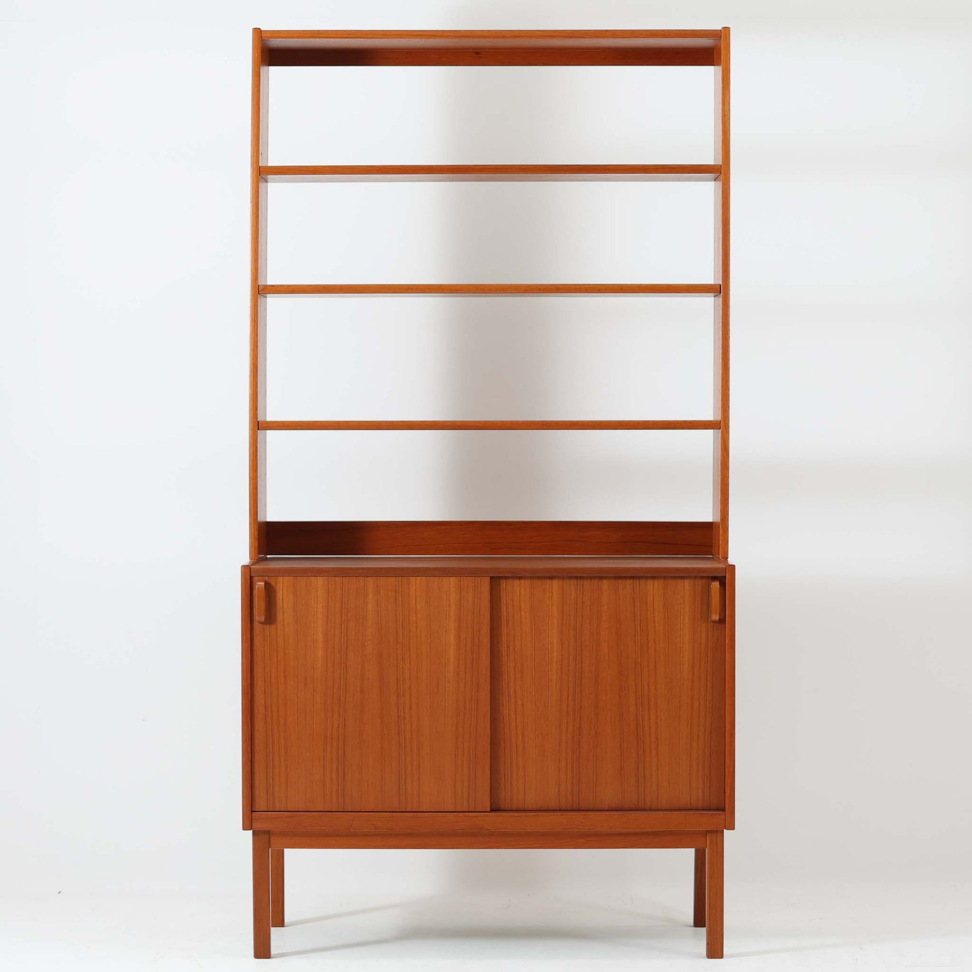 B.フリードホーゲンがデザインし、Bodafors社により製作された北欧ビンテージのディスプレイシェルフです。チーク材の美しい木目を活かした縦に伸びる端正なフレーム構成が印象的で、オープン棚と下部収納を組み合わせた実用性の高いデザインとなっています。上段は抜け感のある棚板構造で、書籍や器、オブジェを軽やかに飾ることができ、下段の引き戸収納は生活感の出やすい物をすっきりと収められます。脚付きによる軽快な佇まいと、経年変化によって深みを増した素材感が、空間に北欧ビンテージならではの落ち着いた存在感を与えます。