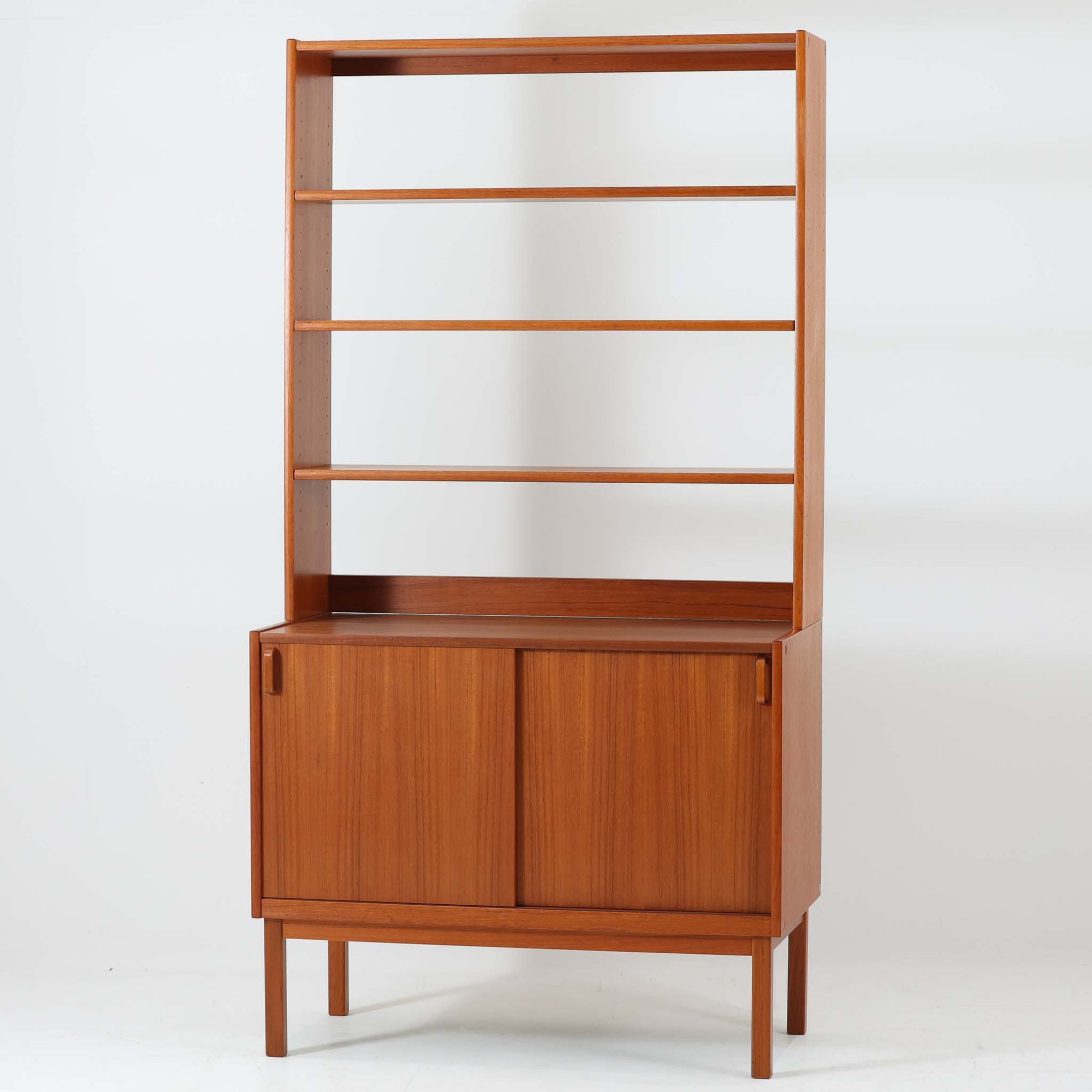 B.フリードホーゲンがデザインし、Bodafors社により製作された北欧ビンテージのディスプレイシェルフです。チーク材の美しい木目を活かした縦に伸びる端正なフレーム構成が印象的で、オープン棚と下部収納を組み合わせた実用性の高いデザインとなっています。上段は抜け感のある棚板構造で、書籍や器、オブジェを軽やかに飾ることができ、下段の引き戸収納は生活感の出やすい物をすっきりと収められます。脚付きによる軽快な佇まいと、経年変化によって深みを増した素材感が、空間に北欧ビンテージならではの落ち着いた存在感を与えます。