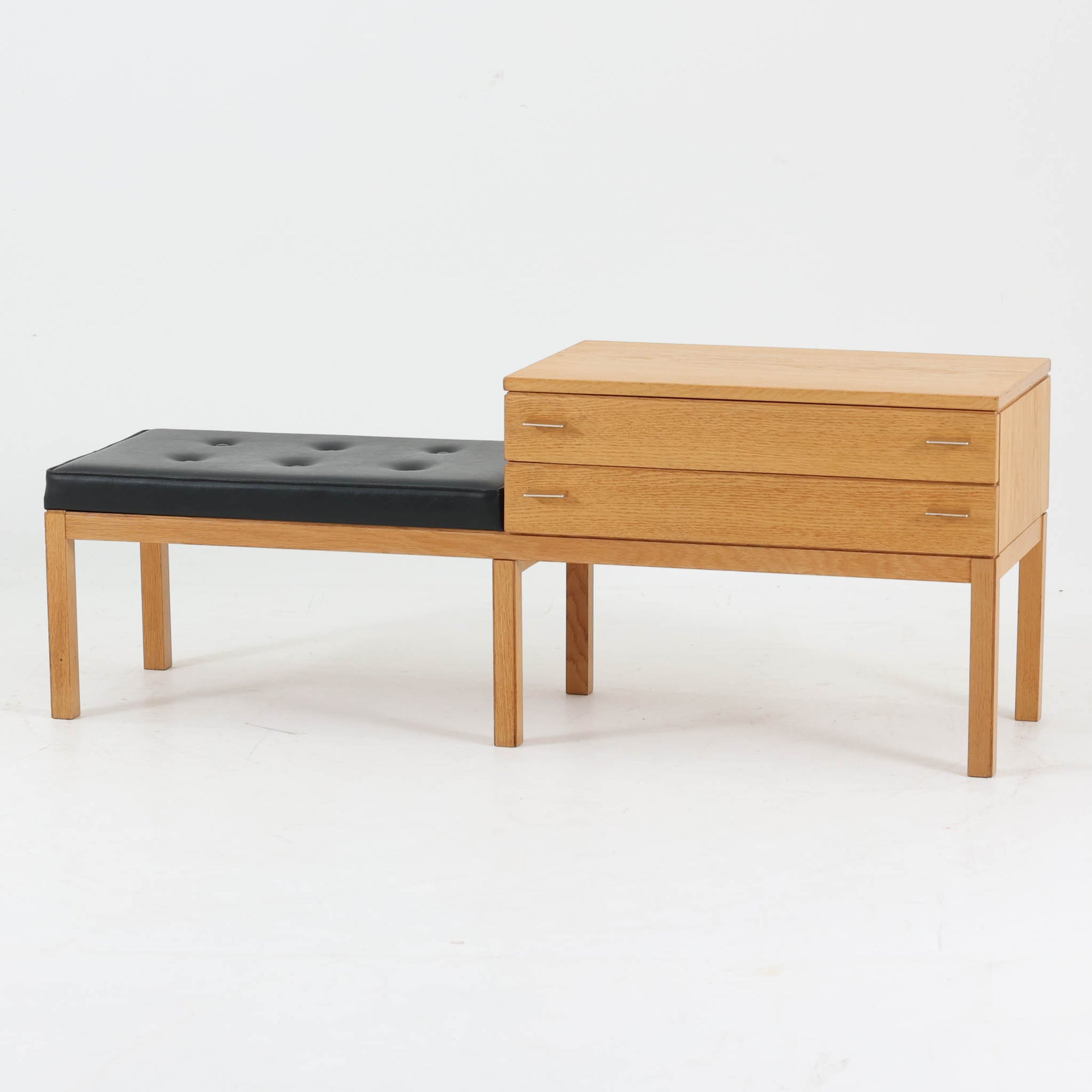 Frosekeが手掛けたTemaシリーズのテレフォンベンチです。オーク材のナチュラルな木目と、ブラックの座面とのコントラストが美しく、空間に引き締まった印象をもたらします。引き出し収納を備えたボックス部とベンチが一体となった構造で、電話台や玄関先での腰掛けとしてはもちろん、小物収納を兼ねた実用的な一台。細身の脚部や控えめなメタルハンドルなど、北欧らしい機能美と軽やかさが魅力です。経年により深みを増した素材感とともに、現代の空間にも自然に溶け込むビンテージならではの存在感をお楽しみいただけます。