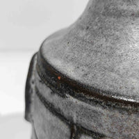 デンマーク製ビンテージテーブルランプです。陶磁器ベースは裾に向かって広がる彫刻的なフォルムを持ち、幾何学的なラインと円形の立体モチーフが規則的に配された、オブジェのような存在感が魅力。マットなグレーを基調とした釉薬に黒の陰影が加わり、手仕事ならではの焼成ムラや表情豊かな質感が際立ちます。リネンシェードから広がるやわらかな灯りが立体意匠を美しく浮かび上がらせ、サイドボードやチェスト、ラウンジ空間のアクセント照明としてもおすすめ。唯一無二の造形美を楽しめる希少なビンテージ品です。