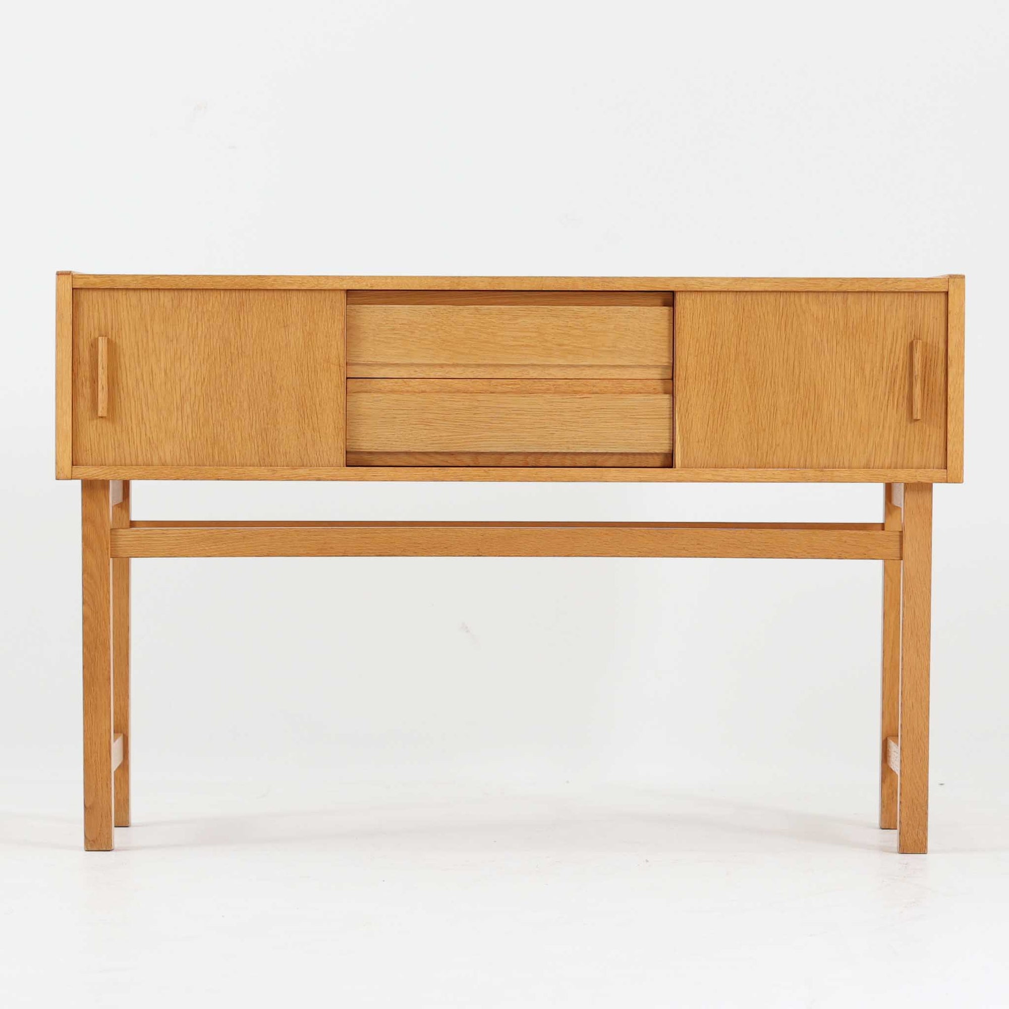 スウェーデン製のローボードです。オーク材ならではの明るく素直な木目と、経年により深みを増した色合いが空間に温もりを添えます。中央に配された引き出しと左右のスライド扉による収納構成は、日用品や書類、小物をすっきり整理できる実用的な設計。細身のフレームと軽やかな脚部、直線を基調とした端正なフォルムが北欧らしい機能美を感じさせます。リビングや寝室での収納兼ディスプレイ台としても使いやすく、ビンテージならではの素材感と丁寧な作りが長く楽しめる一台です。