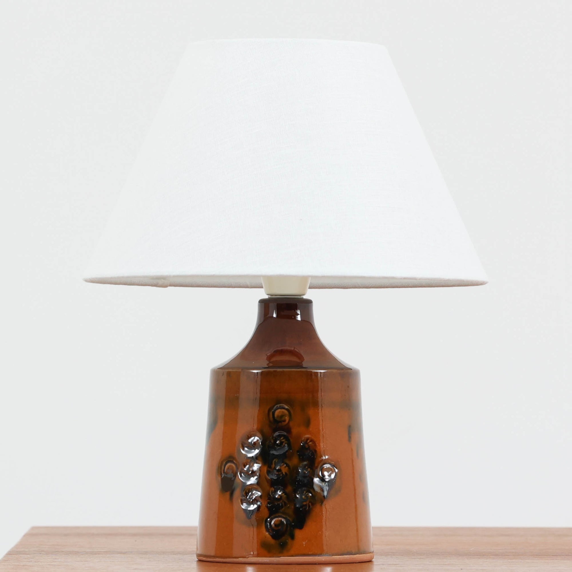 デンマーク製ビンテージテーブルランプです。艶やかな飴色の陶磁器ベースは、裾に向かって穏やかに広がる安定感のあるフォルムに、小花のような立体装飾があしらわれ、素朴さと愛らしさを併せ持つ表情が魅力。焼成によって生まれた黒のにじみや濃淡が一点ごとの個性を際立たせ、手仕事ならではの温もりを感じさせます。リネンシェードからこぼれるやわらかな灯りが、ベッドサイドやチェスト上を穏やかに照らし、北欧らしい落ち着いた空気感を演出。小ぶりながら存在感があり、空間にやさしいアクセントを添えるビンテージ品です。