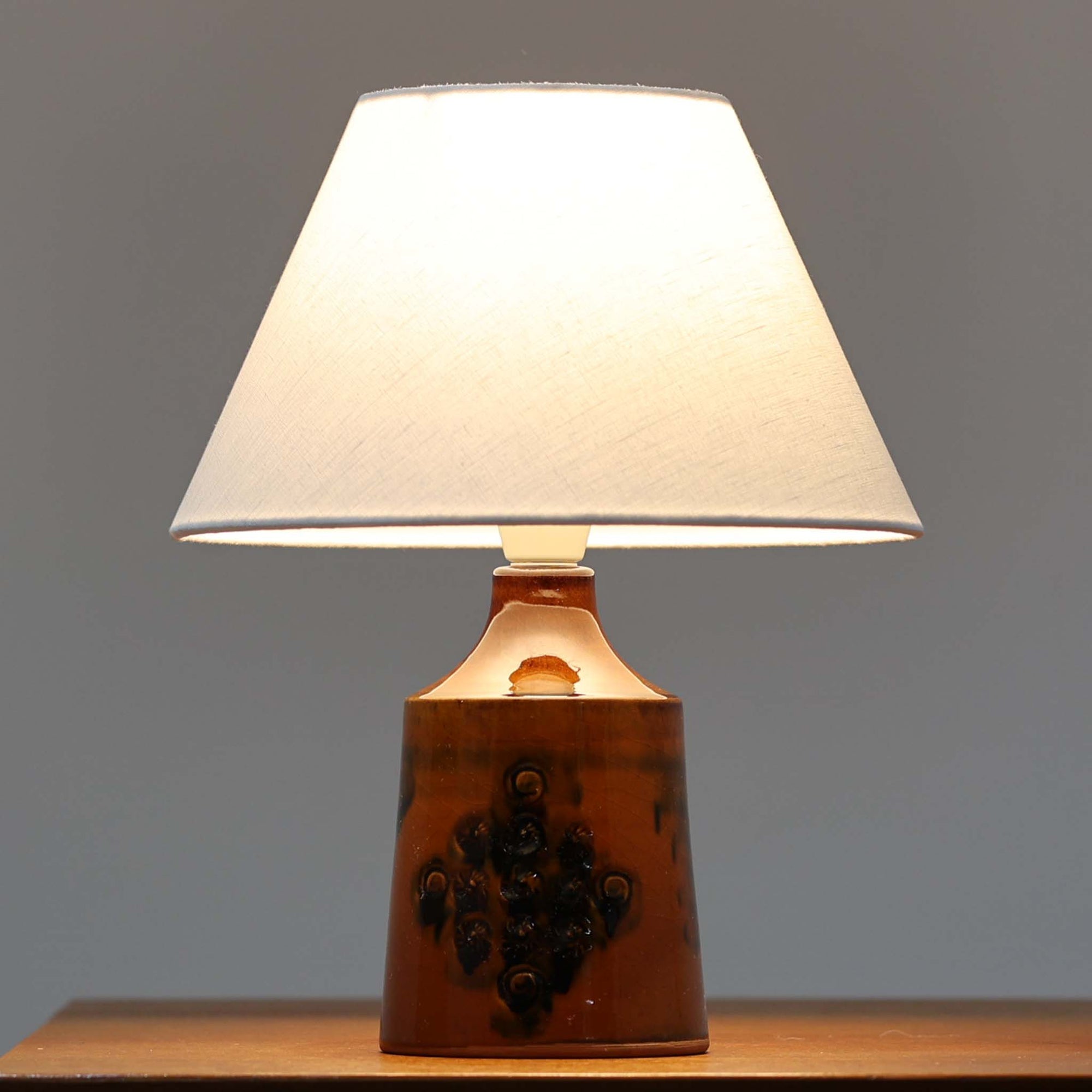 デンマーク製ビンテージテーブルランプです。艶やかな飴色の陶磁器ベースは、裾に向かって穏やかに広がる安定感のあるフォルムに、小花のような立体装飾があしらわれ、素朴さと愛らしさを併せ持つ表情が魅力。焼成によって生まれた黒のにじみや濃淡が一点ごとの個性を際立たせ、手仕事ならではの温もりを感じさせます。リネンシェードからこぼれるやわらかな灯りが、ベッドサイドやチェスト上を穏やかに照らし、北欧らしい落ち着いた空気感を演出。小ぶりながら存在感があり、空間にやさしいアクセントを添えるビンテージ品です。