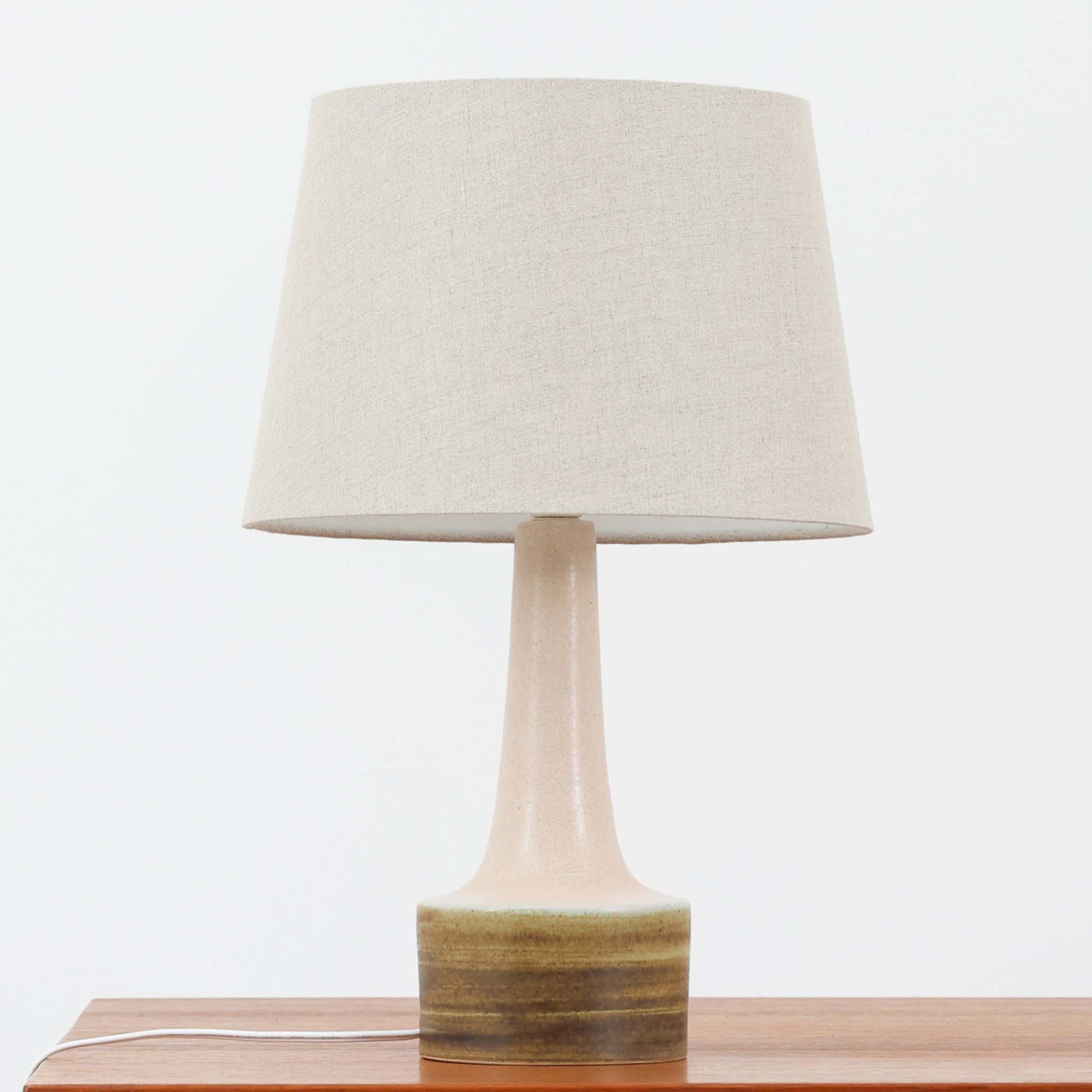 Okela Stonewareによるデンマーク製ビンテージテーブルランプです。すっと立ち上がる細身の陶磁器ベースは、ミニマルで美しいシルエットが印象的。やわらかなアイボリーの陶肌に、裾部分へ重なるブラウンのグラデーション釉が大地を思わせる温かな表情を添えています。装飾を抑えた端正な造形だからこそ、焼成による自然な色の移ろいと素材感が際立つ一台。リネンシェードから広がる穏やかな灯りが、ベッドサイドやサイドボード、ラウンジ空間に静かな上質さをもたらします。北欧らしい素朴な美しさとモダンさを兼ね備えた、長く楽しめるビンテージ品です。