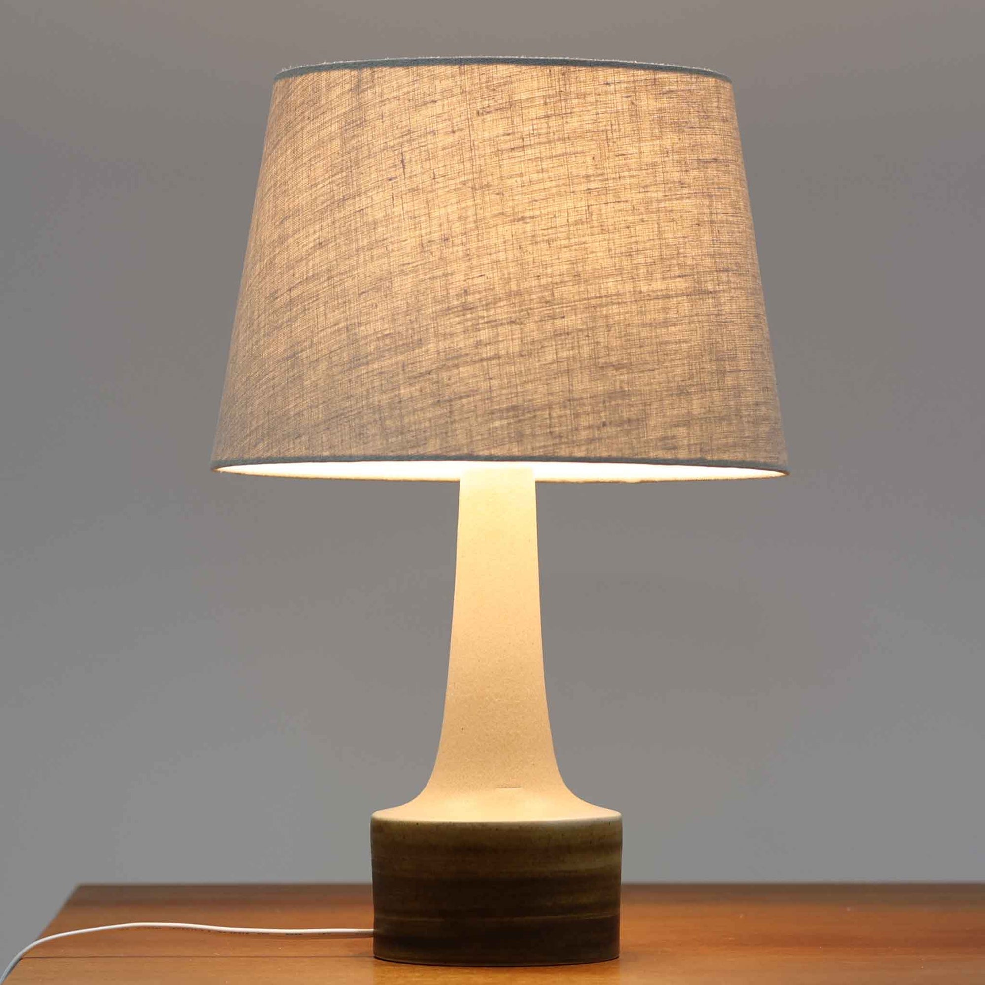 Okela Stonewareによるデンマーク製ビンテージテーブルランプです。すっと立ち上がる細身の陶磁器ベースは、ミニマルで美しいシルエットが印象的。やわらかなアイボリーの陶肌に、裾部分へ重なるブラウンのグラデーション釉が大地を思わせる温かな表情を添えています。装飾を抑えた端正な造形だからこそ、焼成による自然な色の移ろいと素材感が際立つ一台。リネンシェードから広がる穏やかな灯りが、ベッドサイドやサイドボード、ラウンジ空間に静かな上質さをもたらします。北欧らしい素朴な美しさとモダンさを兼ね備えた、長く楽しめるビンテージ品です。