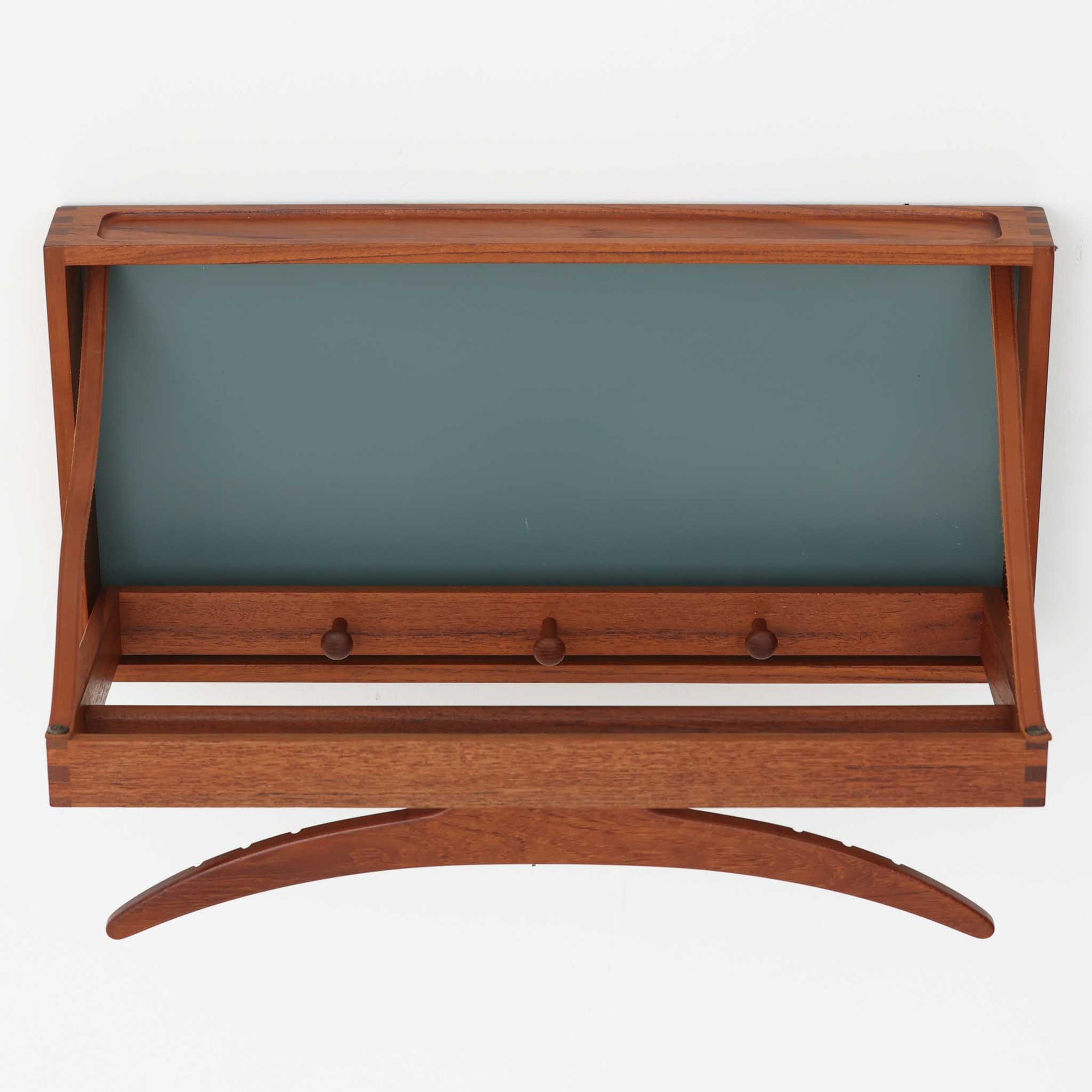 Adam Hoff &amp; Poul Ostergaardがデザインし、Virum Mobelsnickeriにより製作された「The Hanger」ハンガーラックです。チーク材の温かみある木目と、優雅な曲線を描くベースのフォルムが印象的なデザイン。内部にはフックが備えられ、コートや帽子などを掛けて収納できます。フロントのフラップを開くとハンガー部分が現れる機能的な構造で、使用しないときはすっきりとした壁面収納として美しく収まります。革ベルトによる固定や細部の造りにも職人技が感じられる、北欧ビンテージならではの洗練された一品です。