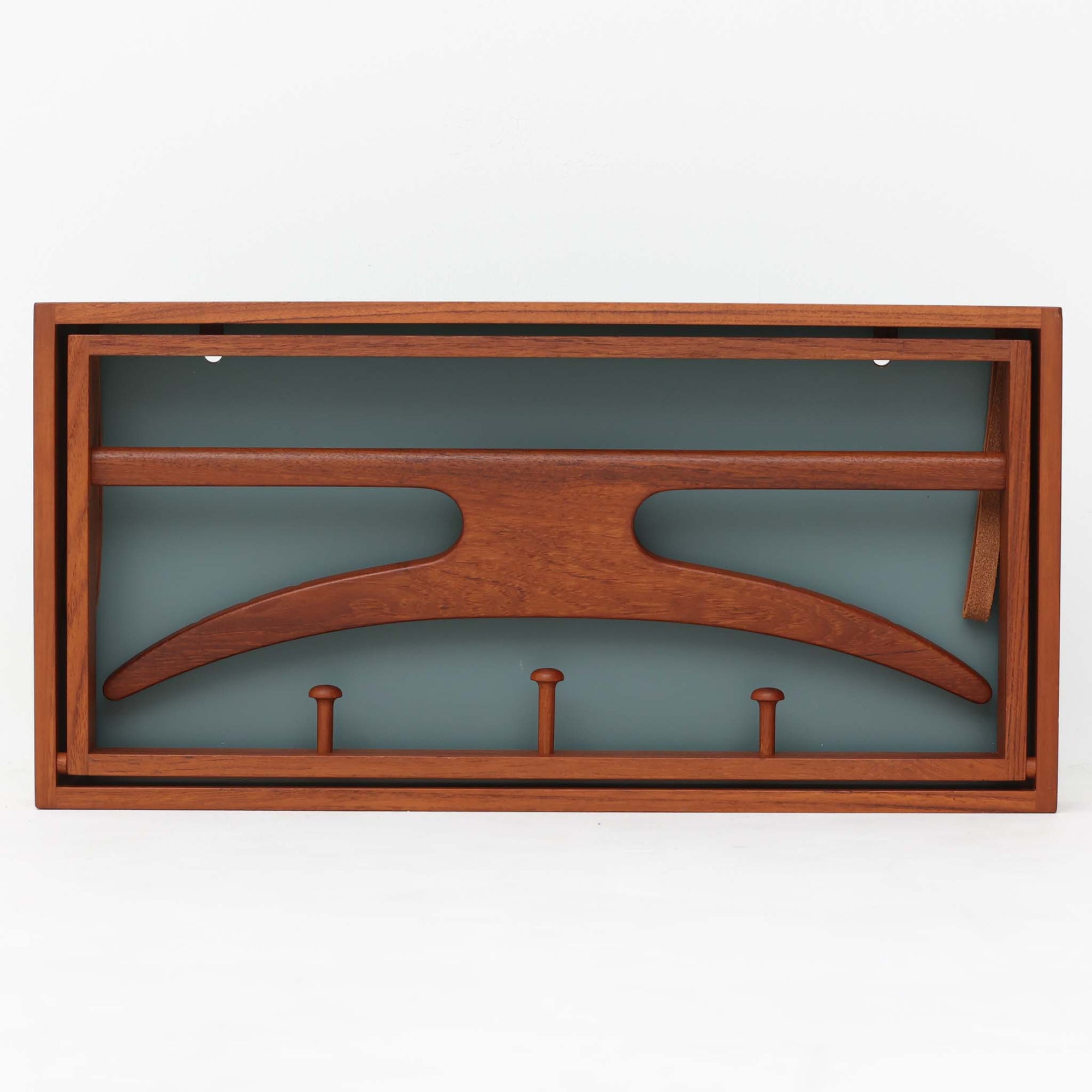 Adam Hoff &amp; Poul Ostergaardがデザインし、Virum Mobelsnickeriにより製作された「The Hanger」ハンガーラックです。チーク材の温かみある木目と、優雅な曲線を描くベースのフォルムが印象的なデザイン。内部にはフックが備えられ、コートや帽子などを掛けて収納できます。フロントのフラップを開くとハンガー部分が現れる機能的な構造で、使用しないときはすっきりとした壁面収納として美しく収まります。革ベルトによる固定や細部の造りにも職人技が感じられる、北欧ビンテージならではの洗練された一品です。