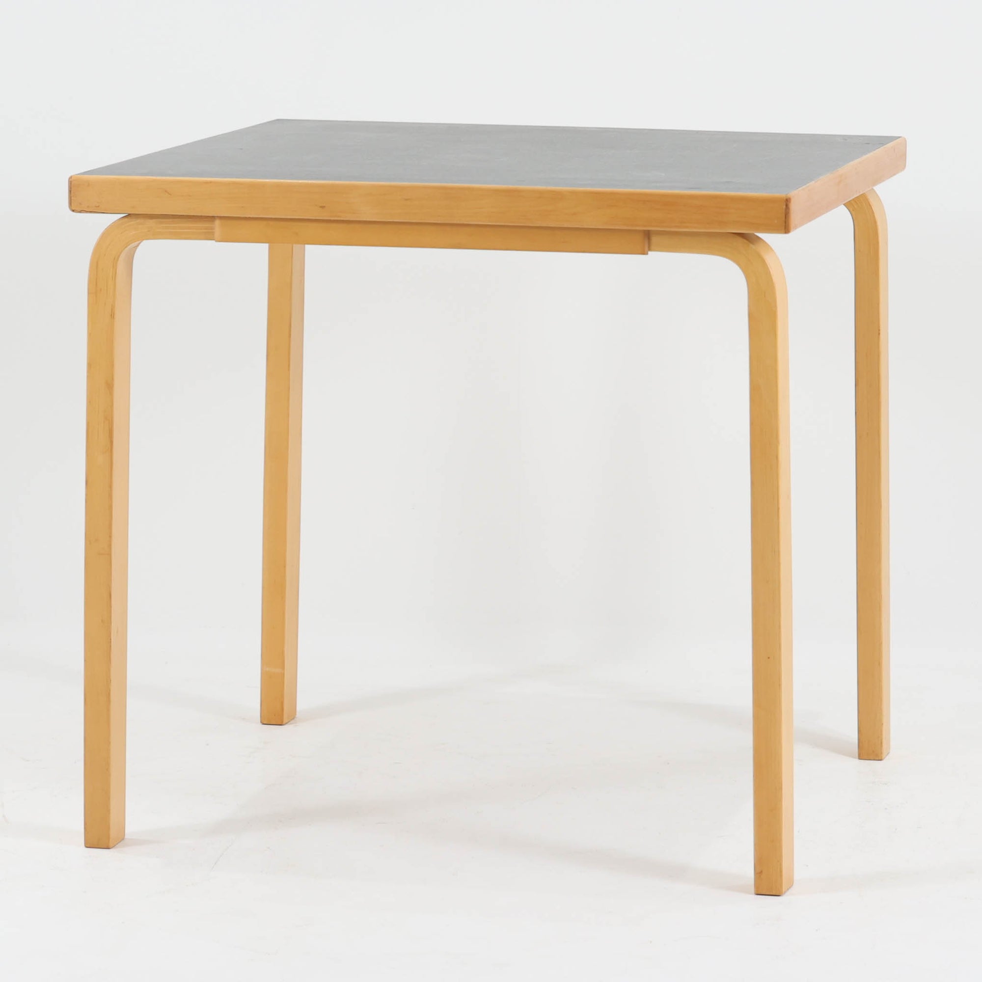 Alvar Aaltoがデザインし、Artekより製作された「81C」ダイニングテーブルです。バーチ材を用いたフレームに、天板には落ち着いた質感のリノリウムを組み合わせたシンプルで機能的なデザインが特徴。脚部にはアアルトを象徴するL字型の曲木構造が採用されており、軽やかで柔らかな印象を与えます。幅75cmの正方形サイズはダイニングはもちろんワークテーブルとしても使いやすく、日常の空間に北欧デザインの美しさと実用性をもたらすビンテージ家具です。本個体は、脚部の5枚レイヤー構造、1973年以降に採用されたプラスネジ、および天板裏の3段構成スタンプの仕様から、1973年～1990年代始めに掛けて製造されたと思われます。