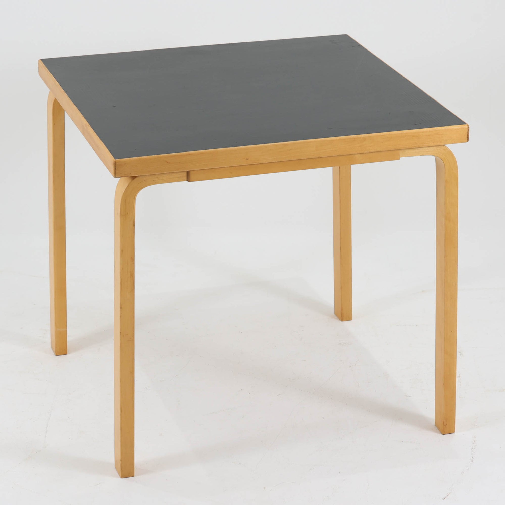 Alvar Aaltoがデザインし、Artekより製作された「81C」ダイニングテーブルです。バーチ材を用いたフレームに、天板には落ち着いた質感のリノリウムを組み合わせたシンプルで機能的なデザインが特徴。脚部にはアアルトを象徴するL字型の曲木構造が採用されており、軽やかで柔らかな印象を与えます。幅75cmの正方形サイズはダイニングはもちろんワークテーブルとしても使いやすく、日常の空間に北欧デザインの美しさと実用性をもたらすビンテージ家具です。本個体は、脚部の5枚レイヤー構造、1973年以降に採用されたプラスネジ、および天板裏の3段構成スタンプの仕様から、1973年～1990年代始めに掛けて製造されたと思われます。