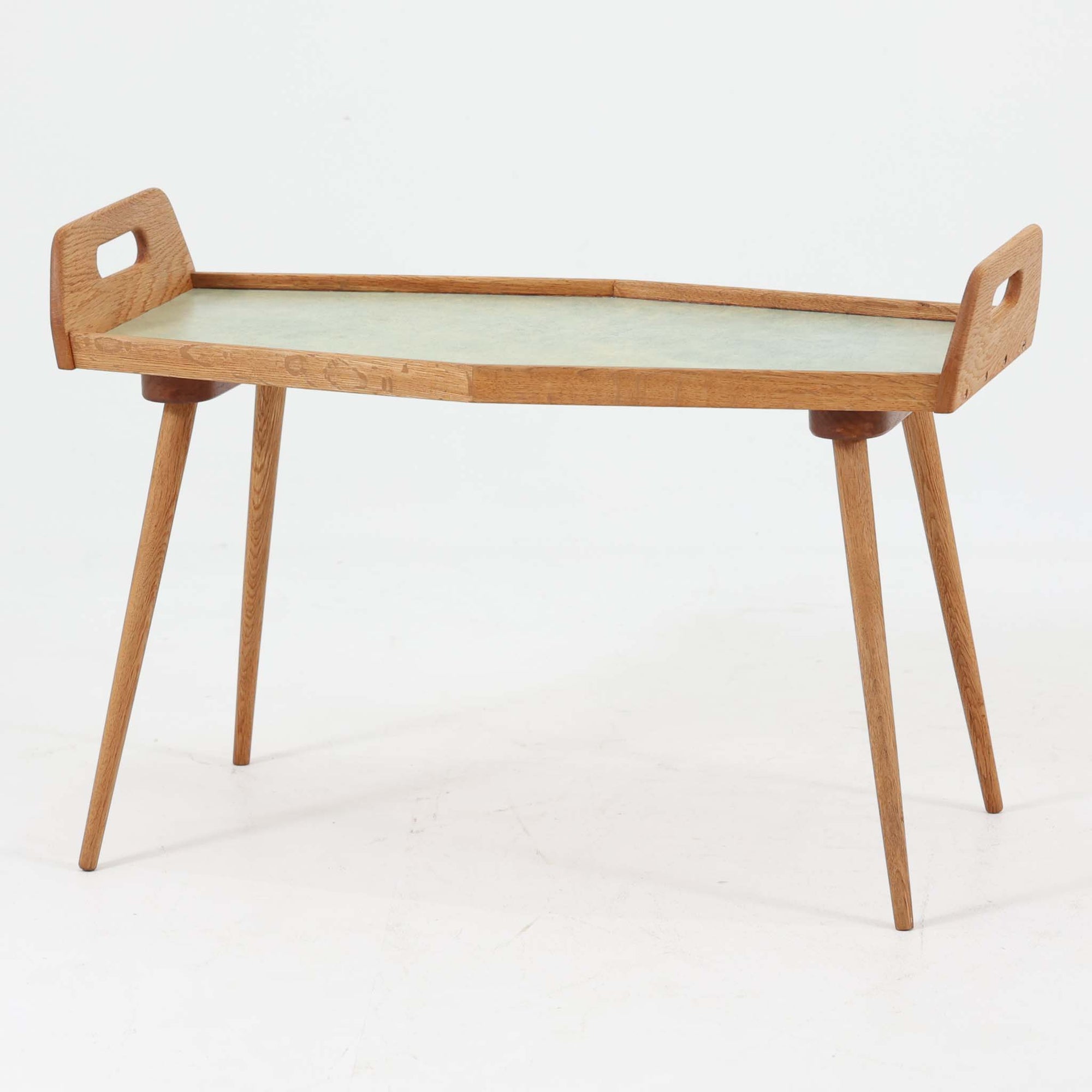 プランターテーブルです。オーク材の温かな木目を活かしたフレームに、淡い色合いのラミネート天板を組み合わせた、北欧らしい軽やかな佇まいが魅力。中央に緩やかな山型を描くユニークな天板形状と、両端の持ち手付きパネル、すらりと伸びるテーパー脚が美しく、置くだけで空間にリズムを生み出します。植物やオブジェを飾るディスプレイ台としてはもちろん、ソファ横のサイドテーブルや玄関先の花台としても使いやすい一台。経年で深みを増した木部の風合いが、ビンテージならではの存在感を引き立てています。