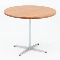 Arne Jacobsenがデザインし、Fritz Hansenが製作した円形ダイニングテーブルです。直径90cmのチーク無垢天板は、豊かな木目と滑らかな円形フォルムが美しく、空間に柔らかな印象をもたらします。シャープなスチール製の一本脚と4本スポークベースが軽快で洗練された佇まいを生み、ヤコブセンらしい機能美が際立つ一台。2～4人で使いやすいサイズ感で、ダイニングテーブルとしてはもちろん、カフェテーブルやラウンジ空間での打ち合わせ用テーブルとしてもおすすめです。中でもチーク無垢材仕様の個体は流通数が少なく珍しく、北欧ビンテージ市場でも希少性の高いモデルとして魅力を放ちます。経年により深みを増した木肌と無駄のない構造が、空間に上質な存在感を添えてくれます。