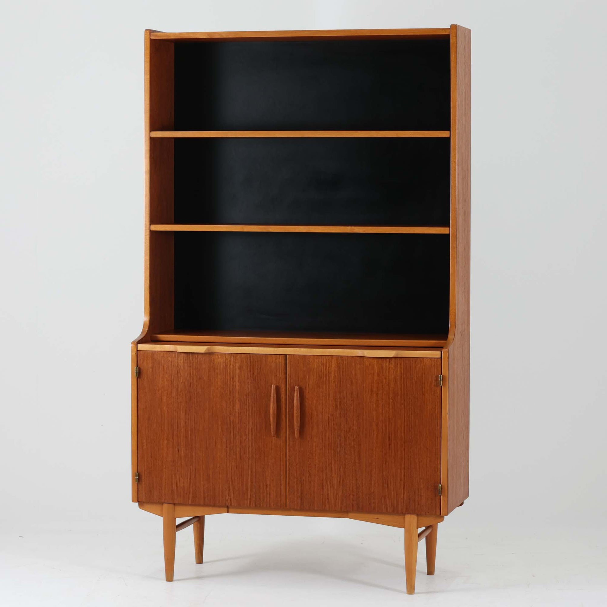 Hugo Troedsが製作したスウェーデン製のディスプレイシェルフです。チーク材の深みある木目とビーチ材の明るい脚部コントラストが美しく、上部のオープン棚と下部キャビネットを組み合わせた実用性の高い構成が魅力。ゆるやかに立ち上がる側板や彫刻的な取っ手、すっきりと伸びる脚部が北欧らしい軽やかな佇まいを演出します。書籍やオブジェ、食器の見せる収納を楽しみながら、下段には生活感の出やすい小物をすっきり収められるため、リビングや書斎、ダイニングボードとしても活躍。経年で増した木肌の艶が空間に上質な存在感を添えるビンテージならではの一品です。