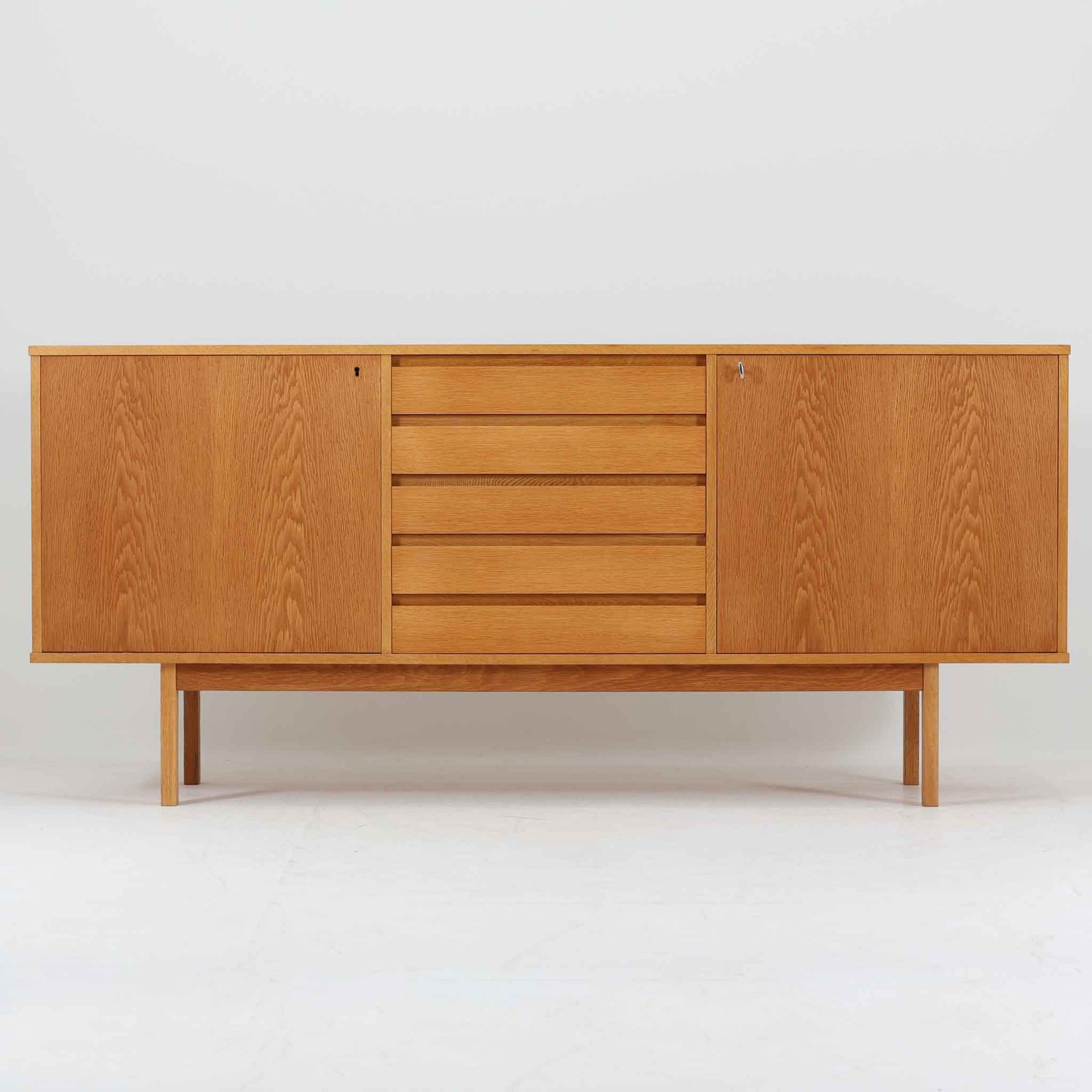 スウェーデン製収納キャビネットです。オーク材ならではの美しい木目と明るく上品な色合いが空間に軽やかな温もりを添え、幅180cmのワイドなサイズが高い収納力を実現します。中央の5段引き出しに加え、左右の扉内には棚板を備え、書類やカトラリー、リビング小物まで用途に応じて整理しやすい実用的な構成。細身の脚部が全体に抜け感を与え、北欧らしいミニマルで端正な佇まいが魅力です。リビングのサイドボードやダイニングでの収納家具としてはもちろん、ショップ什器としても映える、ビンテージならではの希少性と丁寧な木工技術を感じられる一台です。