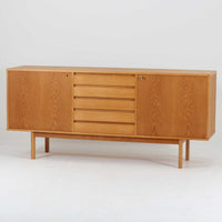 スウェーデン製収納キャビネットです。オーク材ならではの美しい木目と明るく上品な色合いが空間に軽やかな温もりを添え、幅180cmのワイドなサイズが高い収納力を実現します。中央の5段引き出しに加え、左右の扉内には棚板を備え、書類やカトラリー、リビング小物まで用途に応じて整理しやすい実用的な構成。細身の脚部が全体に抜け感を与え、北欧らしいミニマルで端正な佇まいが魅力です。リビングのサイドボードやダイニングでの収納家具としてはもちろん、ショップ什器としても映える、ビンテージならではの希少性と丁寧な木工技術を感じられる一台です。