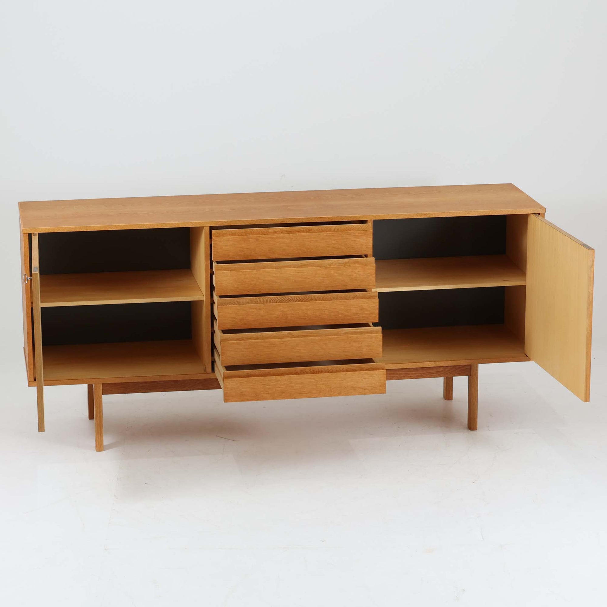 スウェーデン製収納キャビネットです。オーク材ならではの美しい木目と明るく上品な色合いが空間に軽やかな温もりを添え、幅180cmのワイドなサイズが高い収納力を実現します。中央の5段引き出しに加え、左右の扉内には棚板を備え、書類やカトラリー、リビング小物まで用途に応じて整理しやすい実用的な構成。細身の脚部が全体に抜け感を与え、北欧らしいミニマルで端正な佇まいが魅力です。リビングのサイドボードやダイニングでの収納家具としてはもちろん、ショップ什器としても映える、ビンテージならではの希少性と丁寧な木工技術を感じられる一台です。