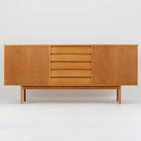 スウェーデン製収納キャビネットです。オーク材ならではの美しい木目と明るく上品な色合いが空間に軽やかな温もりを添え、幅180cmのワイドなサイズが高い収納力を実現します。中央の5段引き出しに加え、左右の扉内には棚板を備え、書類やカトラリー、リビング小物まで用途に応じて整理しやすい実用的な構成。細身の脚部が全体に抜け感を与え、北欧らしいミニマルで端正な佇まいが魅力です。リビングのサイドボードやダイニングでの収納家具としてはもちろん、ショップ什器としても映える、ビンテージならではの希少性と丁寧な木工技術を感じられる一台です。