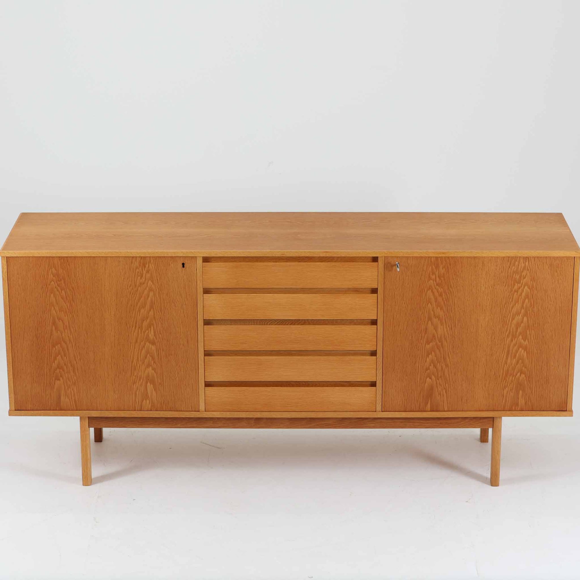 スウェーデン製収納キャビネットです。オーク材ならではの美しい木目と明るく上品な色合いが空間に軽やかな温もりを添え、幅180cmのワイドなサイズが高い収納力を実現します。中央の5段引き出しに加え、左右の扉内には棚板を備え、書類やカトラリー、リビング小物まで用途に応じて整理しやすい実用的な構成。細身の脚部が全体に抜け感を与え、北欧らしいミニマルで端正な佇まいが魅力です。リビングのサイドボードやダイニングでの収納家具としてはもちろん、ショップ什器としても映える、ビンテージならではの希少性と丁寧な木工技術を感じられる一台です。