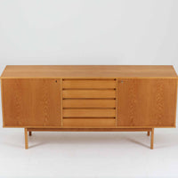 スウェーデン製収納キャビネットです。オーク材ならではの美しい木目と明るく上品な色合いが空間に軽やかな温もりを添え、幅180cmのワイドなサイズが高い収納力を実現します。中央の5段引き出しに加え、左右の扉内には棚板を備え、書類やカトラリー、リビング小物まで用途に応じて整理しやすい実用的な構成。細身の脚部が全体に抜け感を与え、北欧らしいミニマルで端正な佇まいが魅力です。リビングのサイドボードやダイニングでの収納家具としてはもちろん、ショップ什器としても映える、ビンテージならではの希少性と丁寧な木工技術を感じられる一台です。