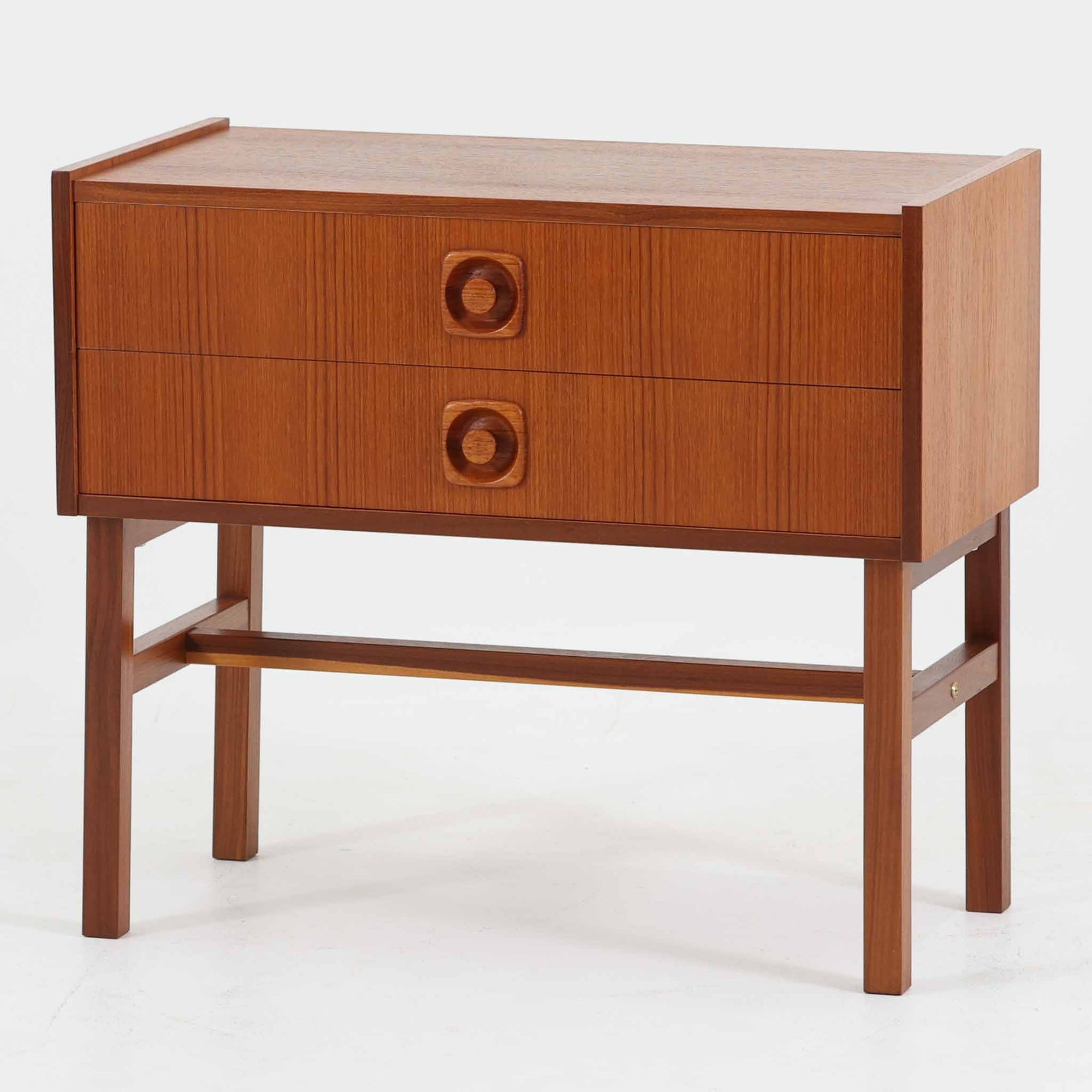スウェーデン製チーク材2段チェストです。幅58cmの取り入れやすいサイズに、縦方向の美しい木目を活かした前板と印象的な円形の削り出し取っ手が映える、北欧らしい端正な一台。2杯の引き出しは衣類や日用品、小物をすっきり整理しやすく、ソファ横のサイドチェストやベッドサイドのナイトテーブルとしても活躍します。細身の脚部と貫が全体に軽やかな印象を与え、経年で深みを増したチークの艶やかな木肌が空間に上質な温もりを添えます。限られた空間にも置きやすく、ビンテージならではの丁寧な造りと希少性を楽しめる収納家具です。