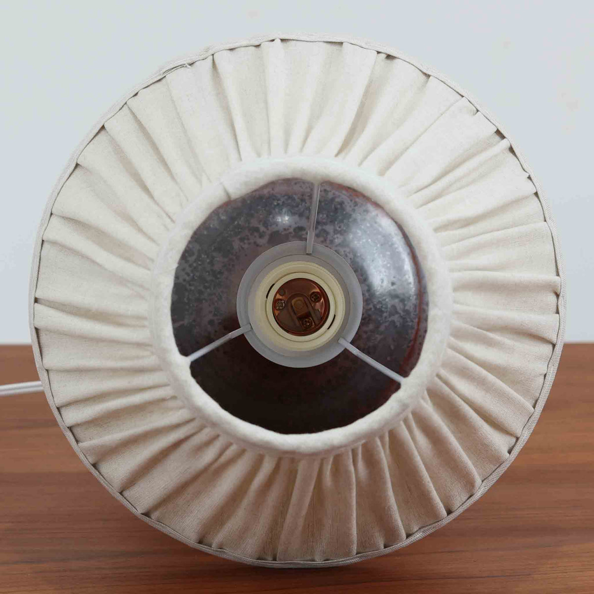 デンマークのDesireeによる陶磁器製のテーブルランプ。深みのある赤褐色を基調に、黒やグレーが複雑に溶け合う釉薬の表情が印象的な一台です。裾に向かって大きく広がる丸みを帯びたフォルムと、すっきりと伸びる首元のバランスが美しく、陶磁器ならではの重厚感と優雅さを兼ね備えています。細かなプリーツを施したリネンシェードは、点灯すると柔らかな光を透過し、温かみのある落ち着いた雰囲気を演出します。消灯時にもオブジェのような存在感があり、リビングのサイドボードや寝室のベッドサイドなど、さまざまな空間におすすめです。釉薬の豊かな色彩と質感に、北欧ビンテージ陶器ならではの魅力が感じられます。